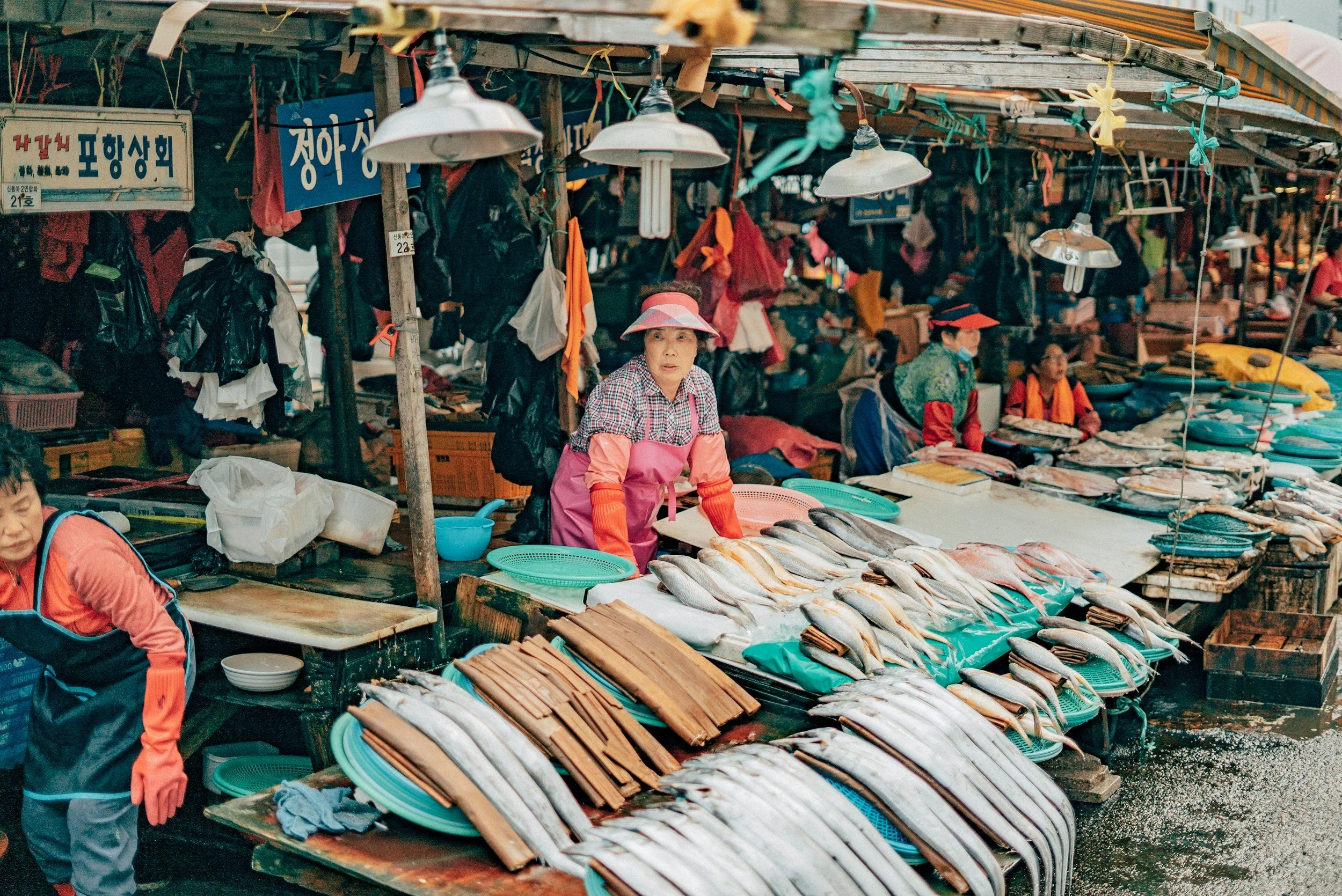 Fish market with vendors selling fish on wet tables, some fish are arranged on trays, in an outdoor covered area with signs in Korean, hanging lights, and market goods in the background.