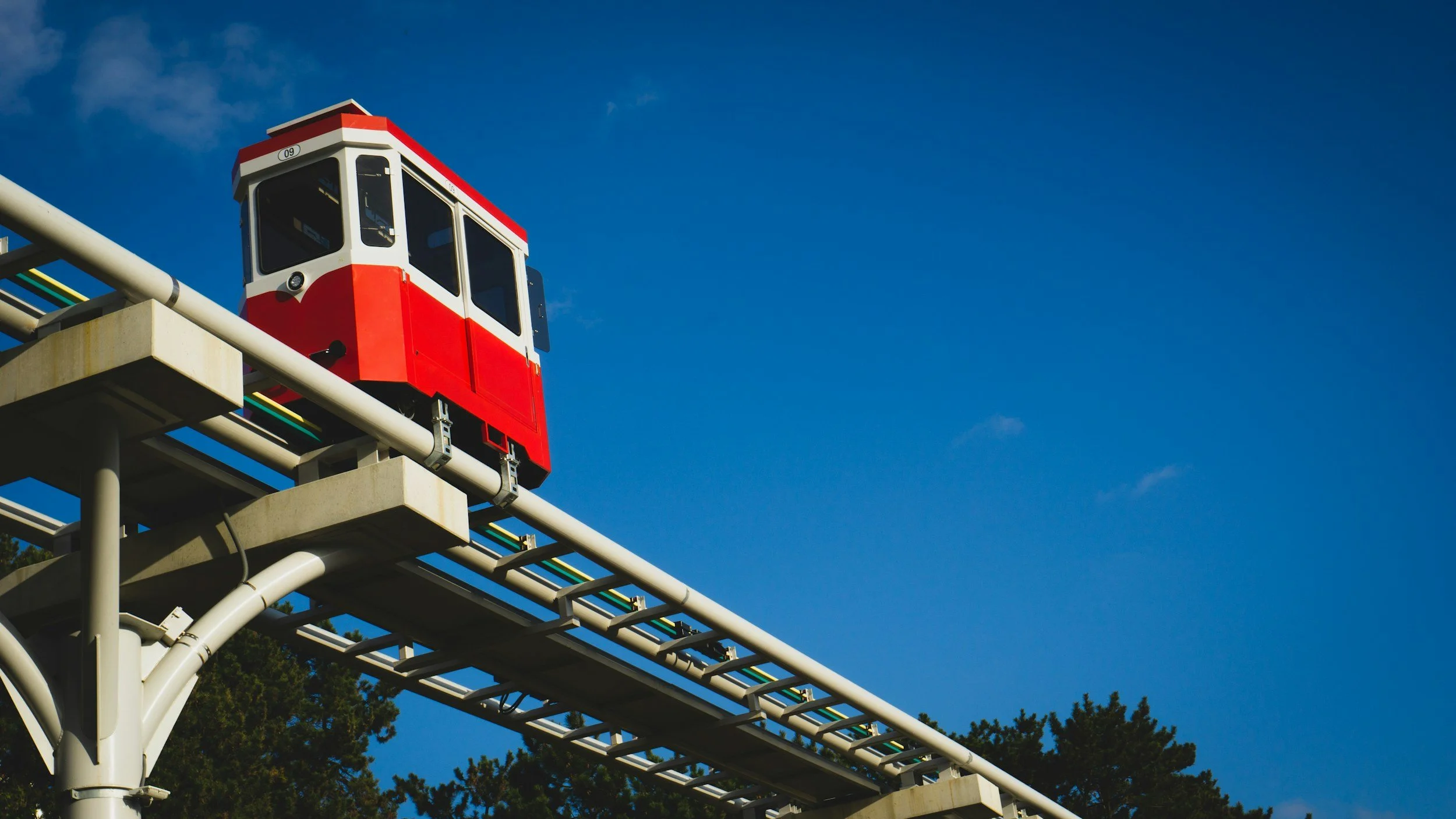 A red and white monorail train on an elevated track against a blue sky with some clouds and trees in the background.