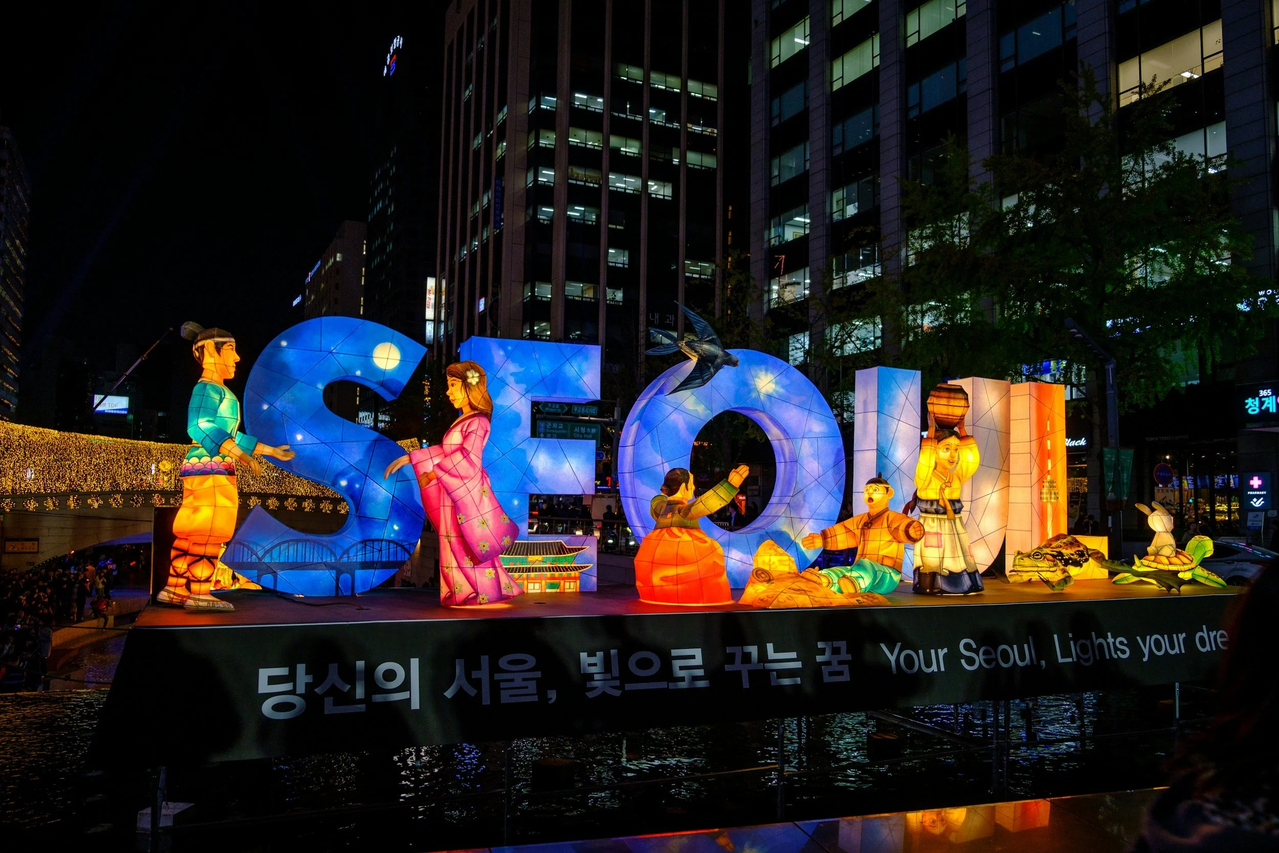 Nighttime city scene with illuminated lantern display spelling 'SEO' and depicting traditional Korean figures and symbols, with Korean and English text on a banner reading 'Your Seoul, Lights your dream'.