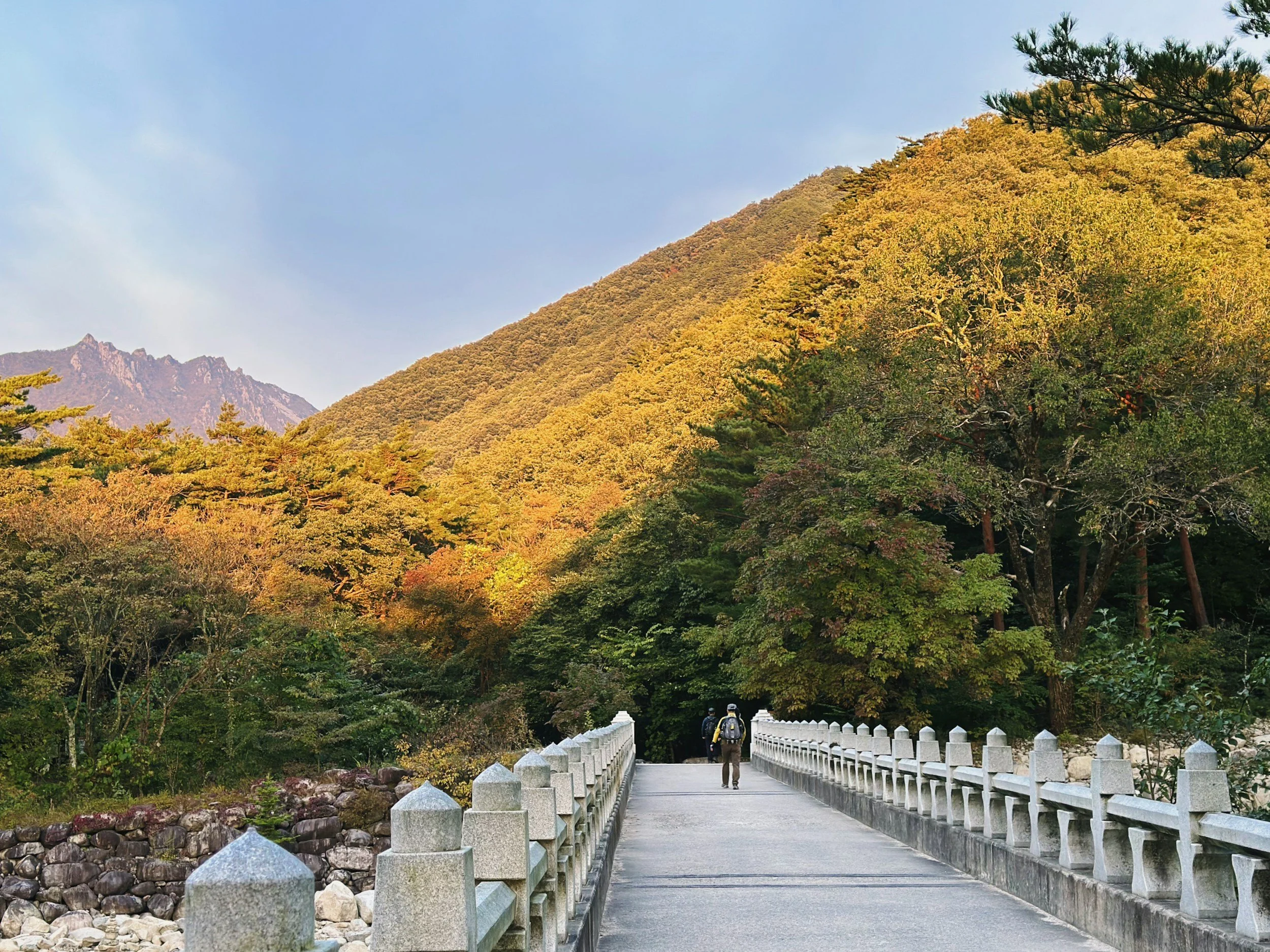 A scenic mountain landscape with autumn-colored trees and a bridge with two hikers walking across.