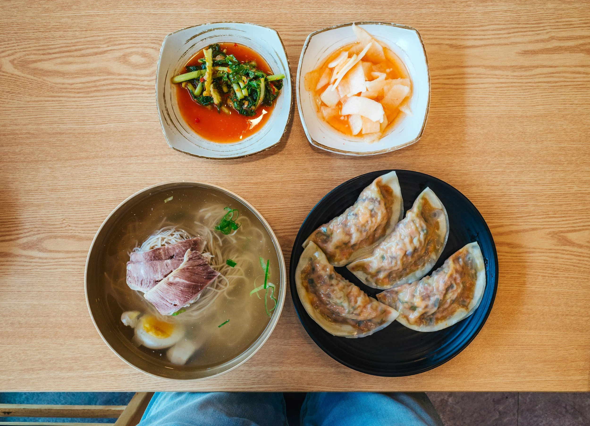 A meal on a wooden table including a bowl of ramen with sliced pork and a soft-boiled egg, a black plate of four dumplings, and two small bowls with dipping sauces and pickled vegetables.