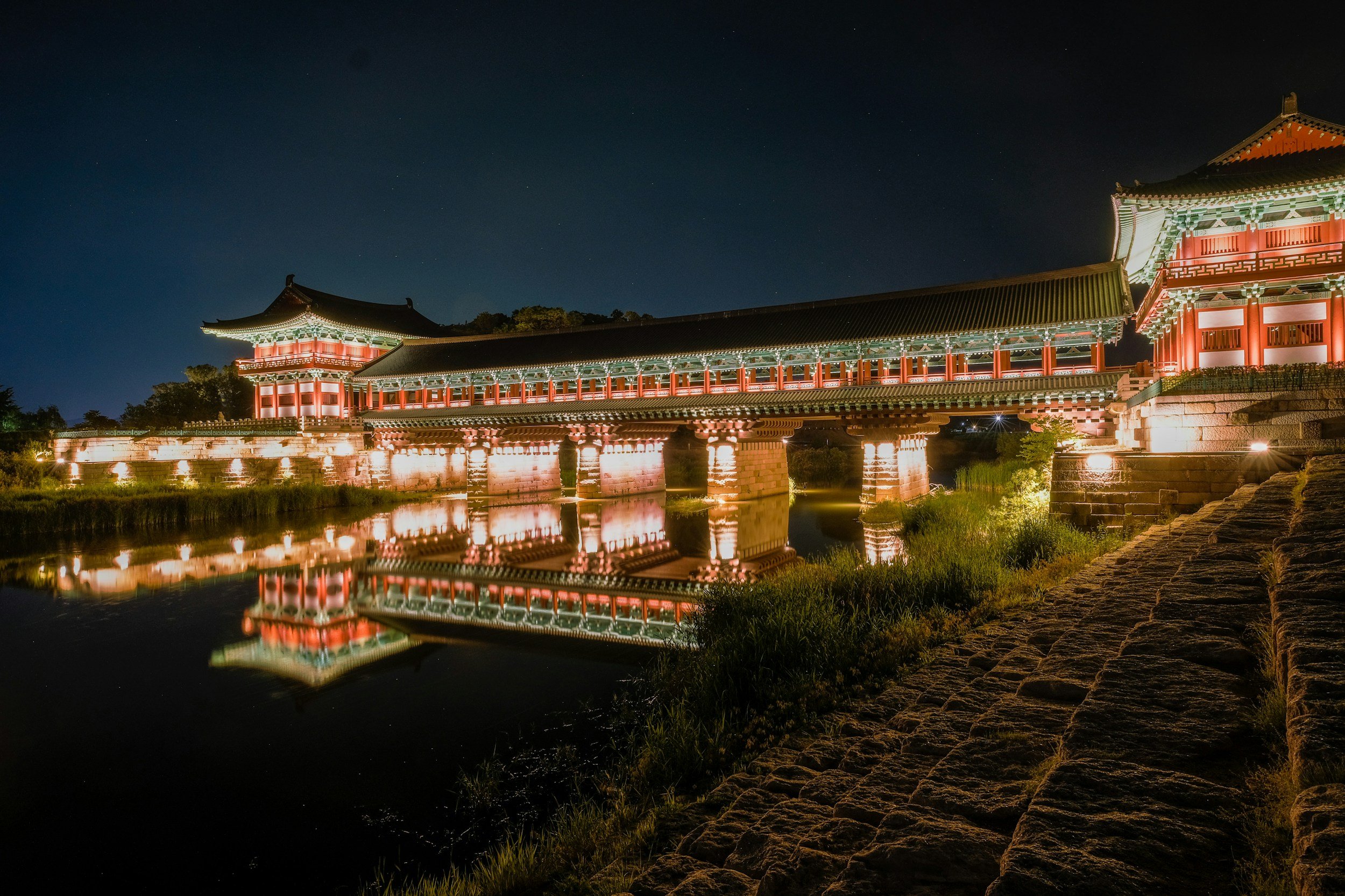 Night view of a traditional Asian bridge with red and green details, illuminated and reflected in a river, with a dark starry sky in the background.
