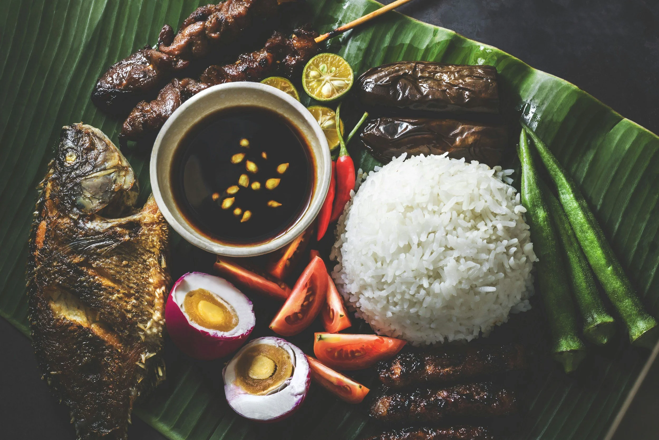 A traditional Malaysian Nasi Lemak plate with rice, fried fish, satay skewers, boiled eggs, eggplant, sliced tomatoes, cucumber, lime, and a small bowl of soy sauce with chili seeds, on a green banana leaf.
