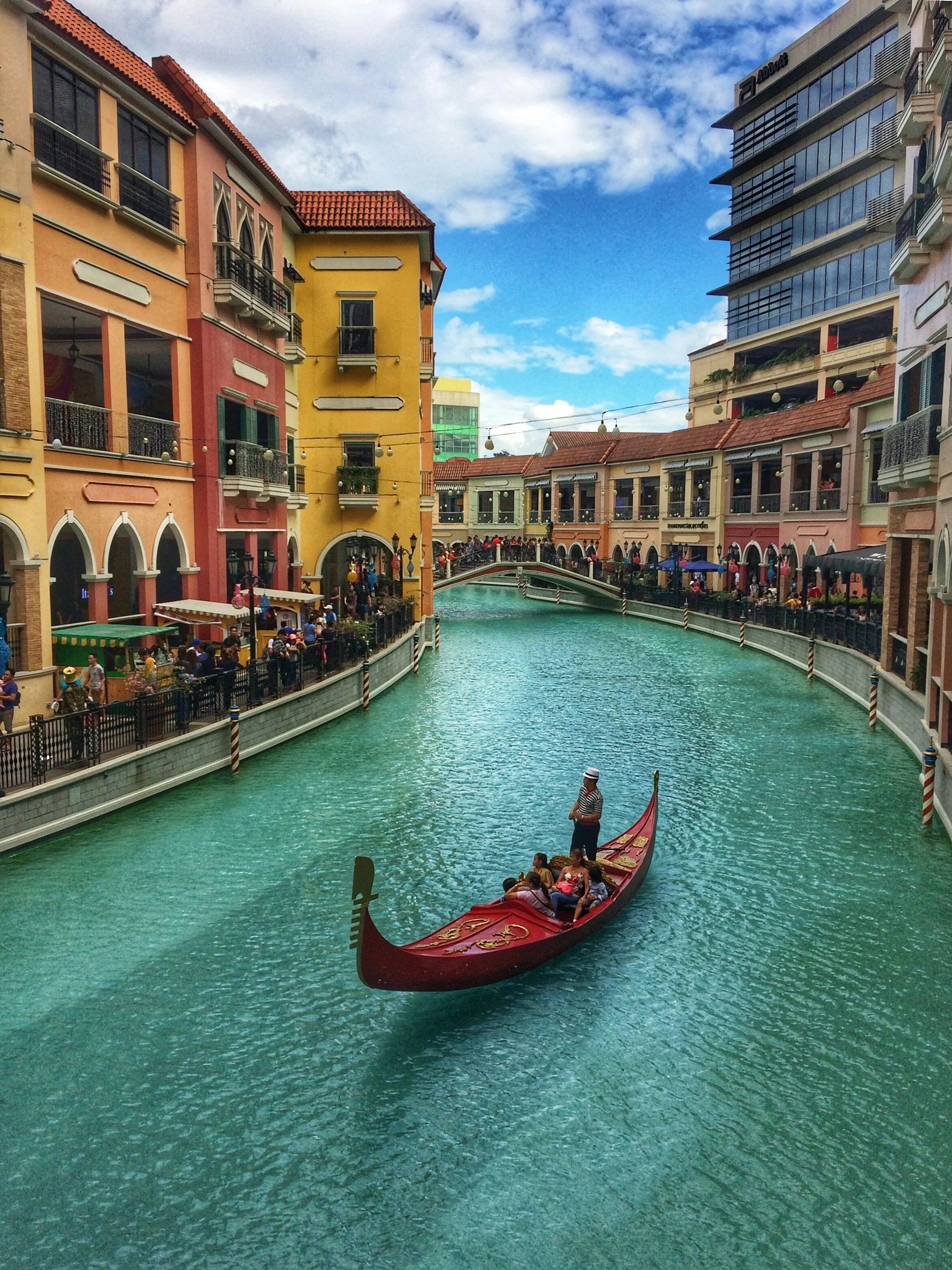 Gondola ride on a canal in a colorful shopping and dining district with European-style buildings and a crowd of people walking along the sides.