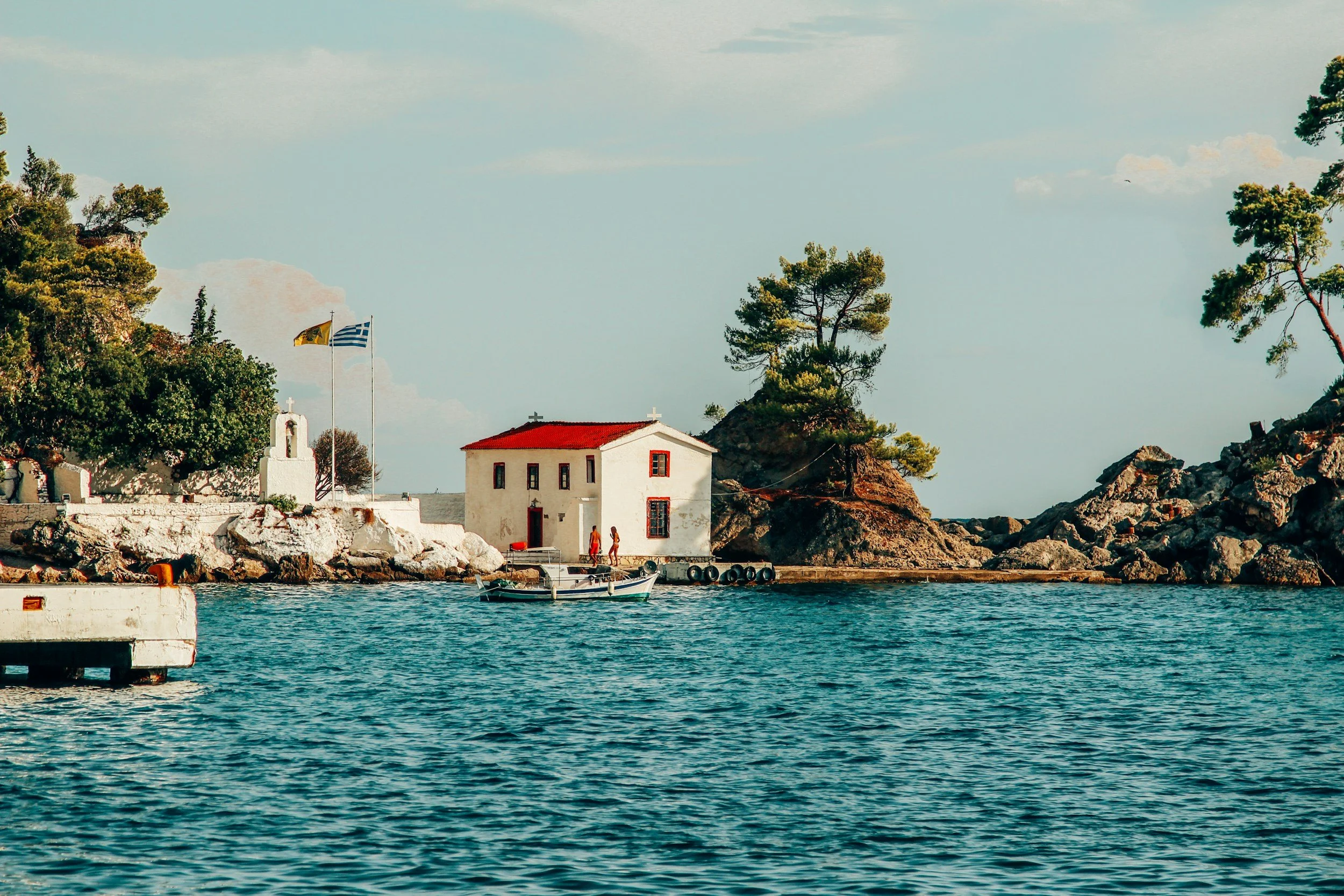 Small white house with red roof near rocky shoreline, trees, boat in water, Greek flag, and chapel cross in background.
