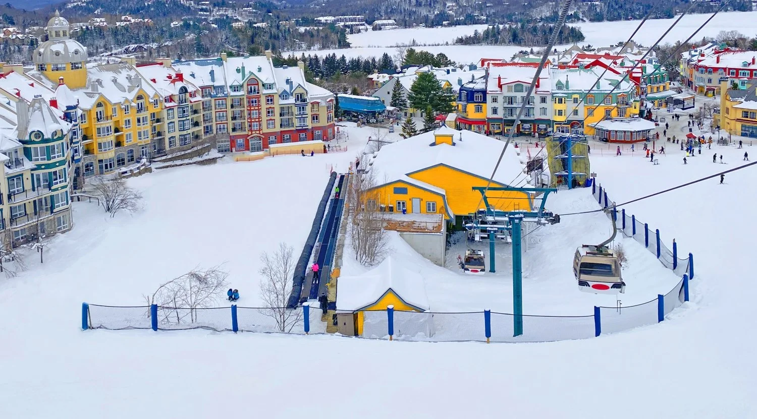 A ski resort town covered in snow with colorful buildings, a ski lift with chairs, and people skiing and walking.