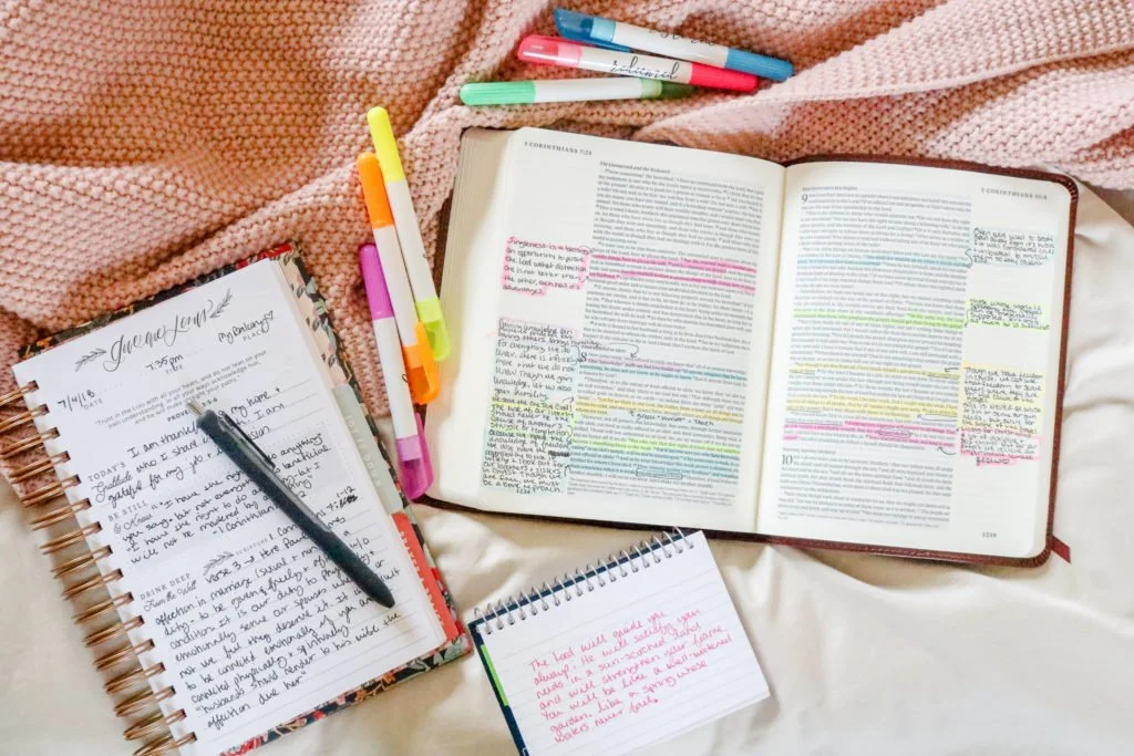 Open Bible with highlighted and handwritten notes, a spiral notebook with handwritten notes, a planner, and colorful pens on a pink and beige blanket.