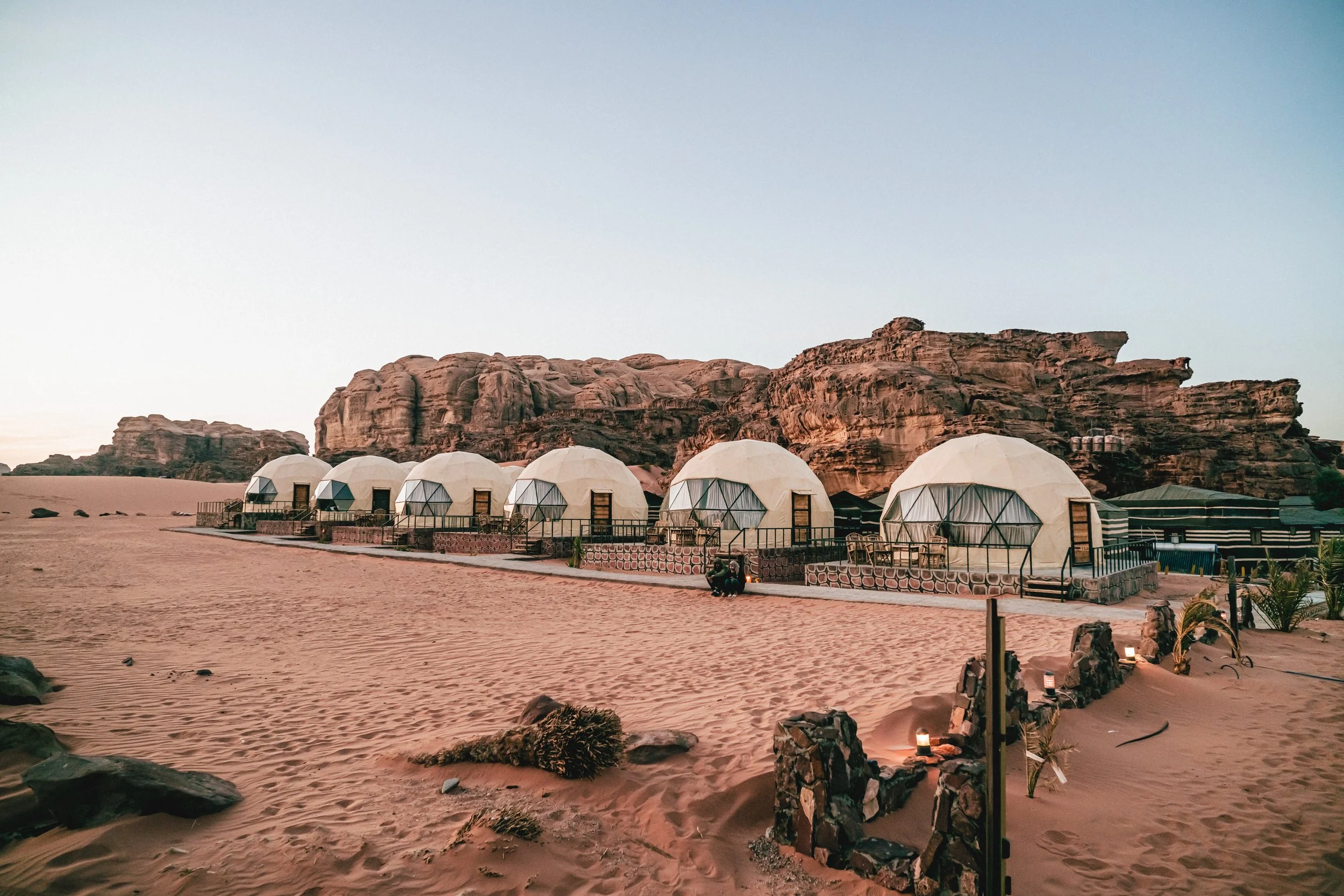 Six white geodesic-style dome tents are set up on a sandy desert landscape with rocky hills in the background, with a person sitting near the tents.