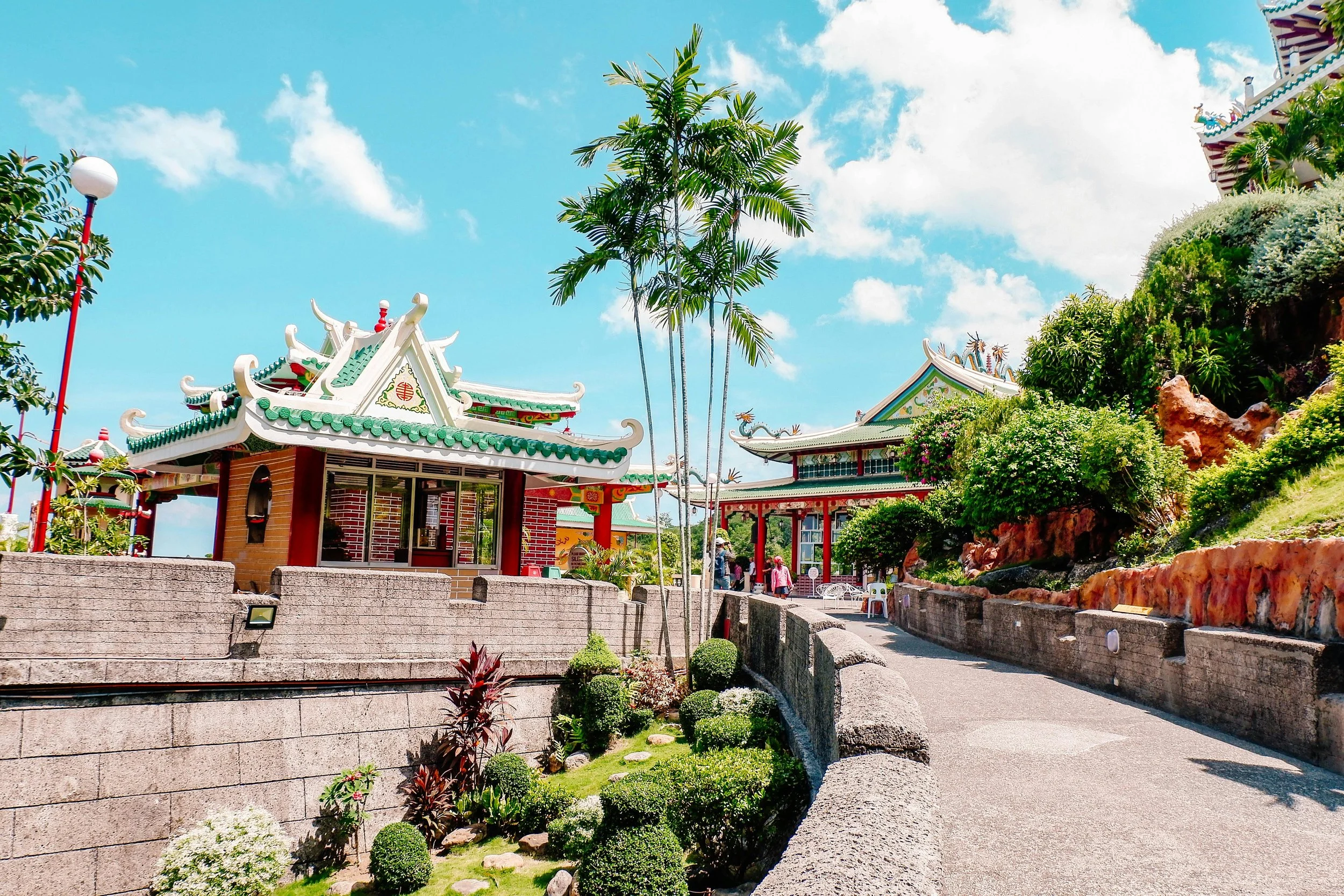 A colorful outdoor Asian-themed park area with traditional pagoda-style structures, palm trees, and lush greenery under a bright blue sky with scattered clouds.