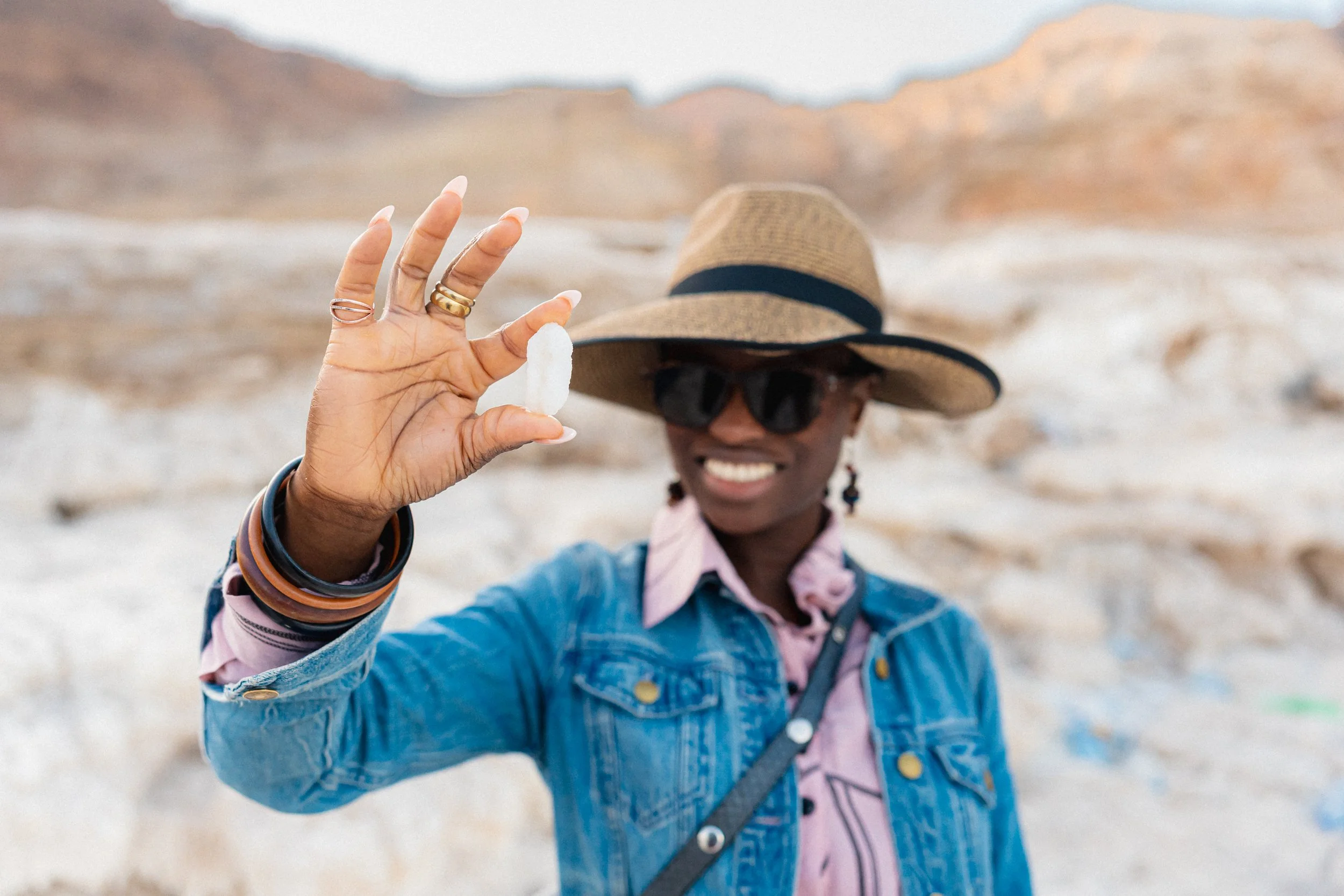 Smiling woman in sunglasses and a large sun hat holding a small white object in her hand, standing outdoors in a rocky desert landscape.