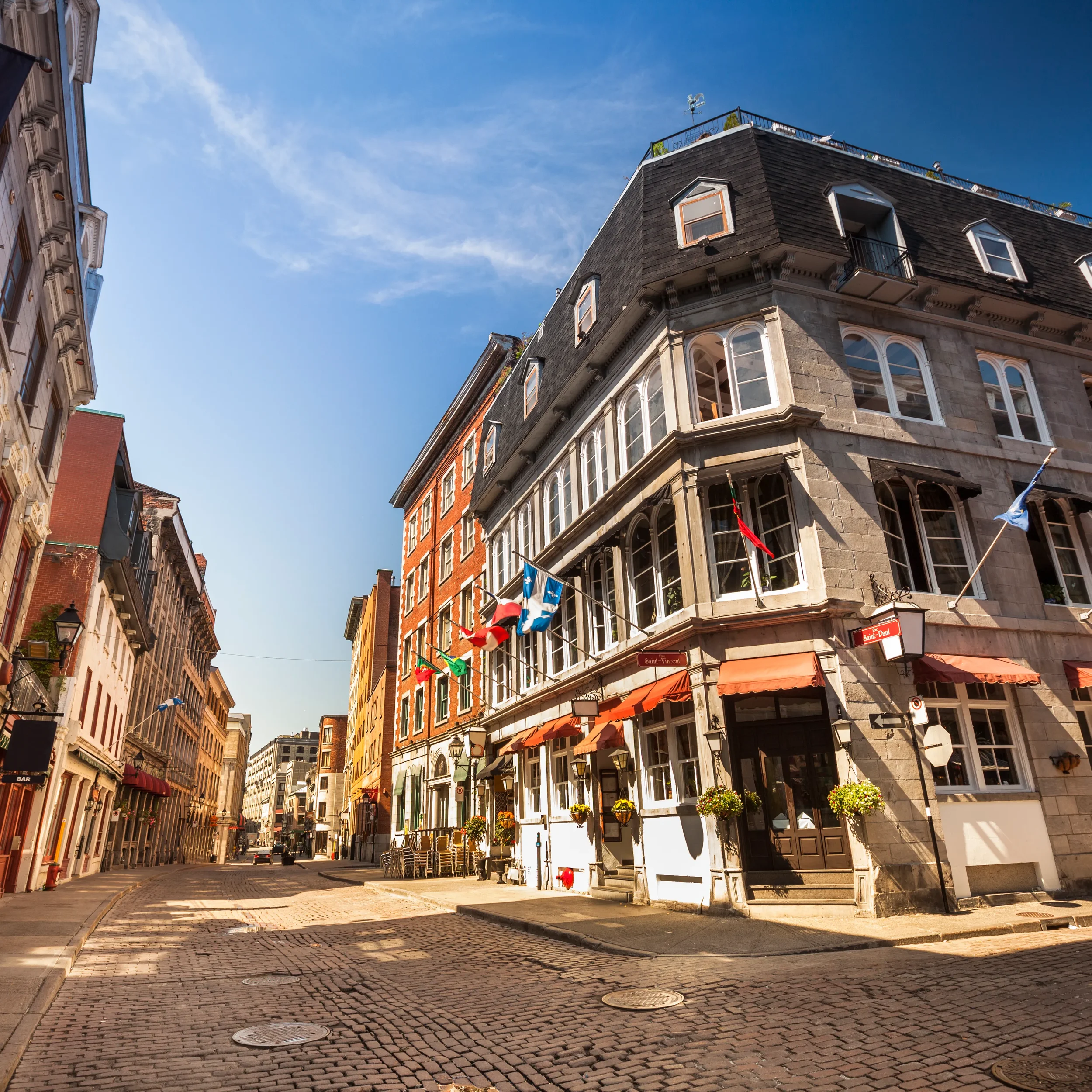 A quiet European city street lined with historic buildings, topped with flags, under a clear blue sky.