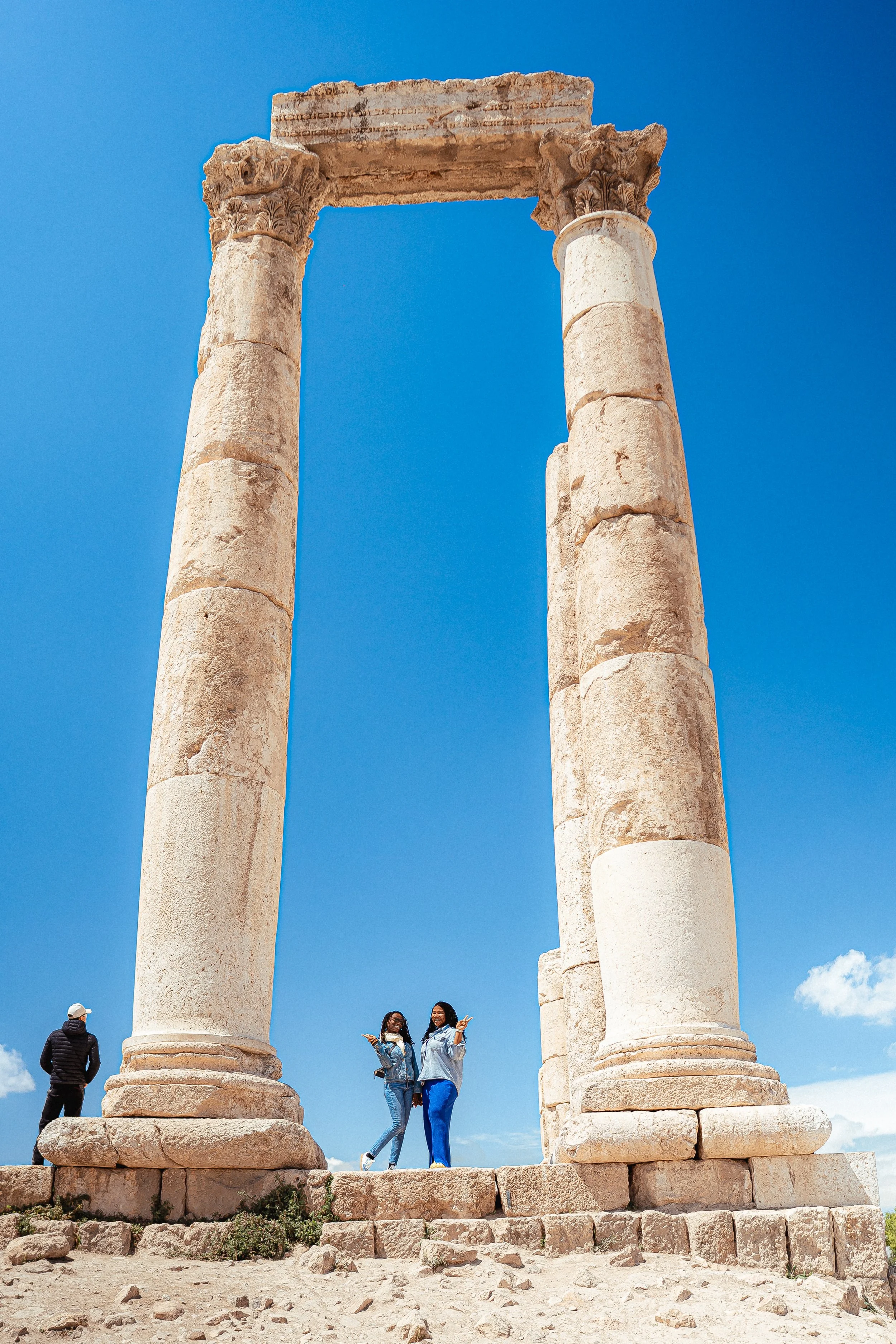 Two women standing under ancient stone columns against a bright blue sky, with a third person walking nearby, at a historical site or ruins.
