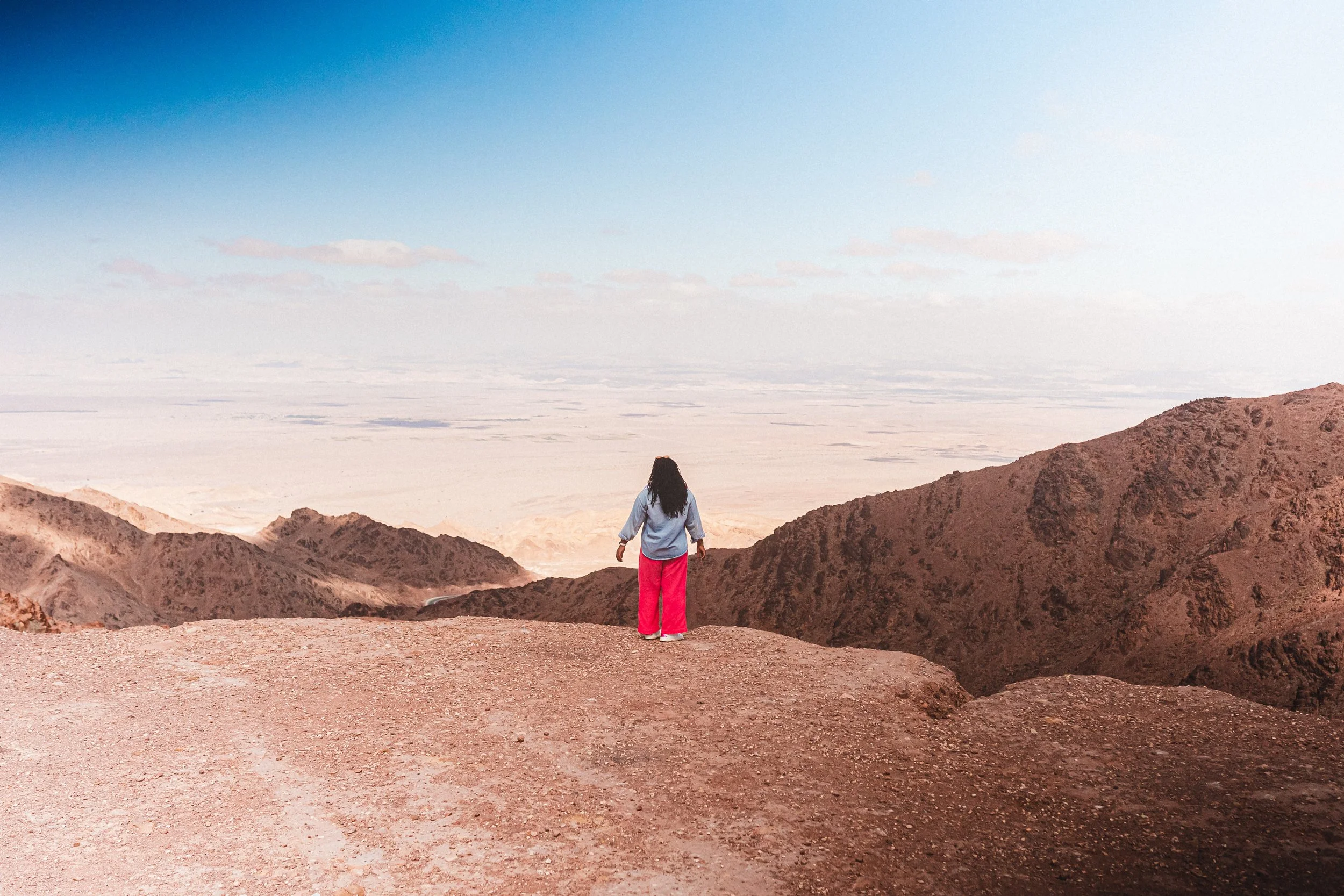 A person standing on rocky terrain overlooking mountains and a desert landscape with a light blue sky.