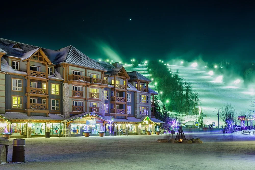 A large multi-story winter resort building decorated with holiday lights, with a ski slope illuminated by green lights and fog in the background at night.