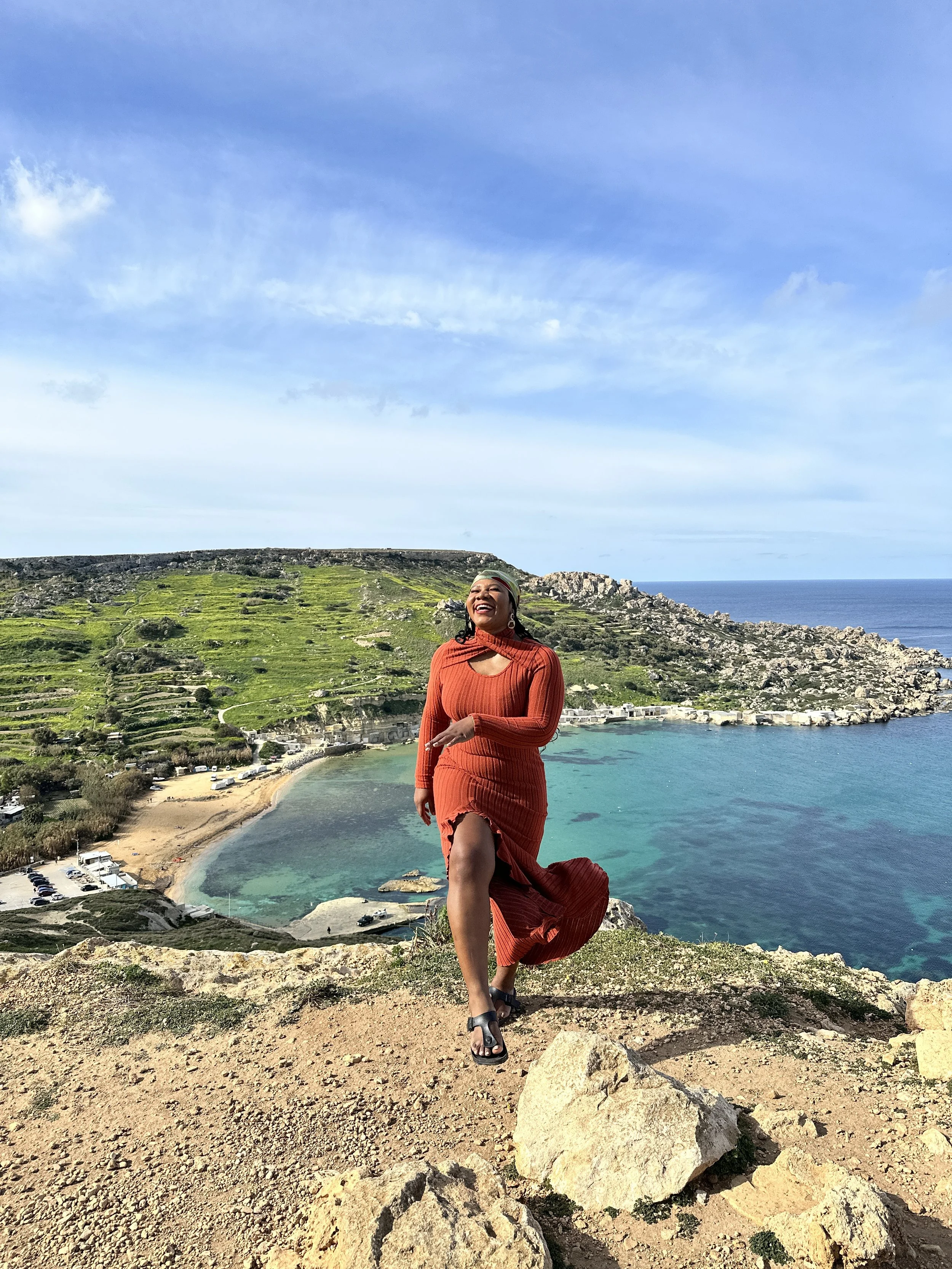 A woman in a red dress standing on a rocky hilltop overlooking a coastal bay with turquoise water, green hillside, and a blue sky with clouds.
