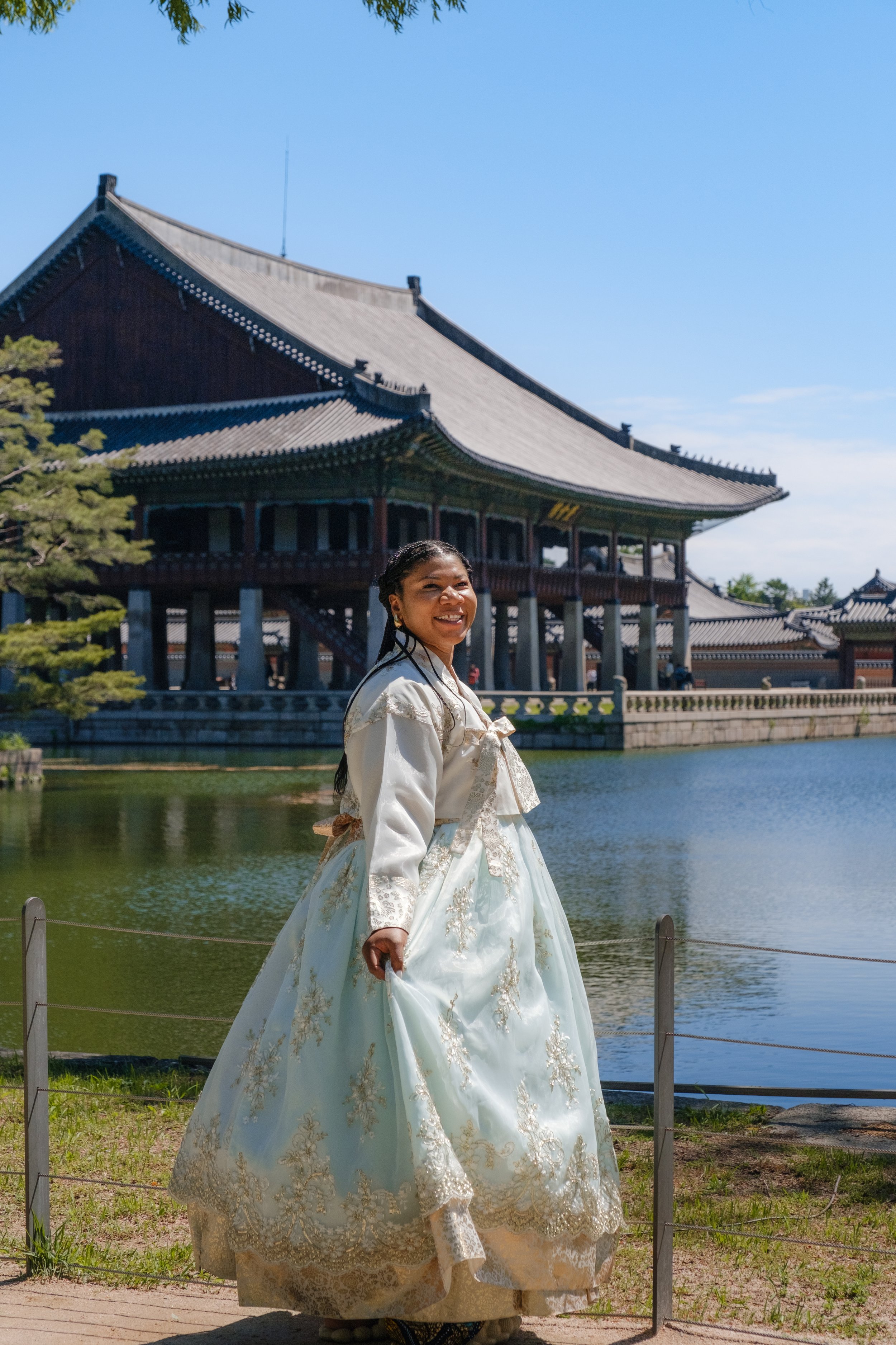 A young woman in traditional Korean hanbok attire, smiling, standing outdoors near a body of water with a historic Korean palace building in the background.