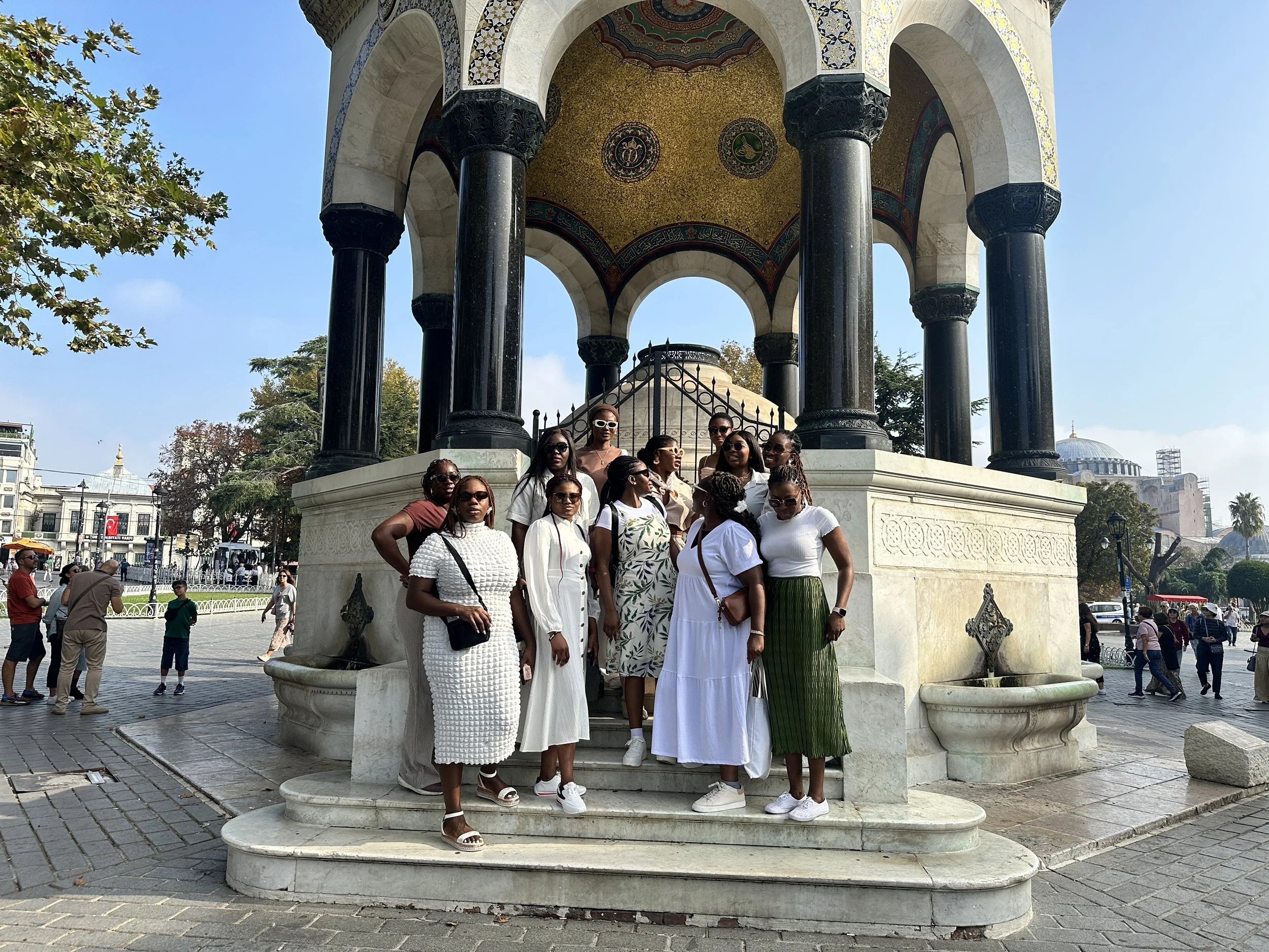 A group of women in casual summer clothing and sunglasses posing on the steps of an ornate, historic gazebo in an outdoor plaza on a sunny day.