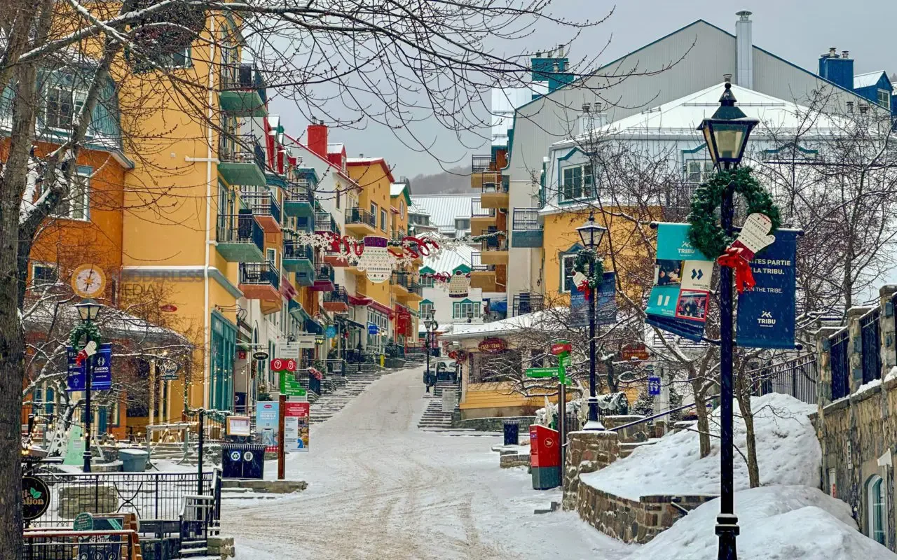Snow-covered street lined with colorful buildings and decorated with holiday wreaths and garlands, with street lamps and signs visible.