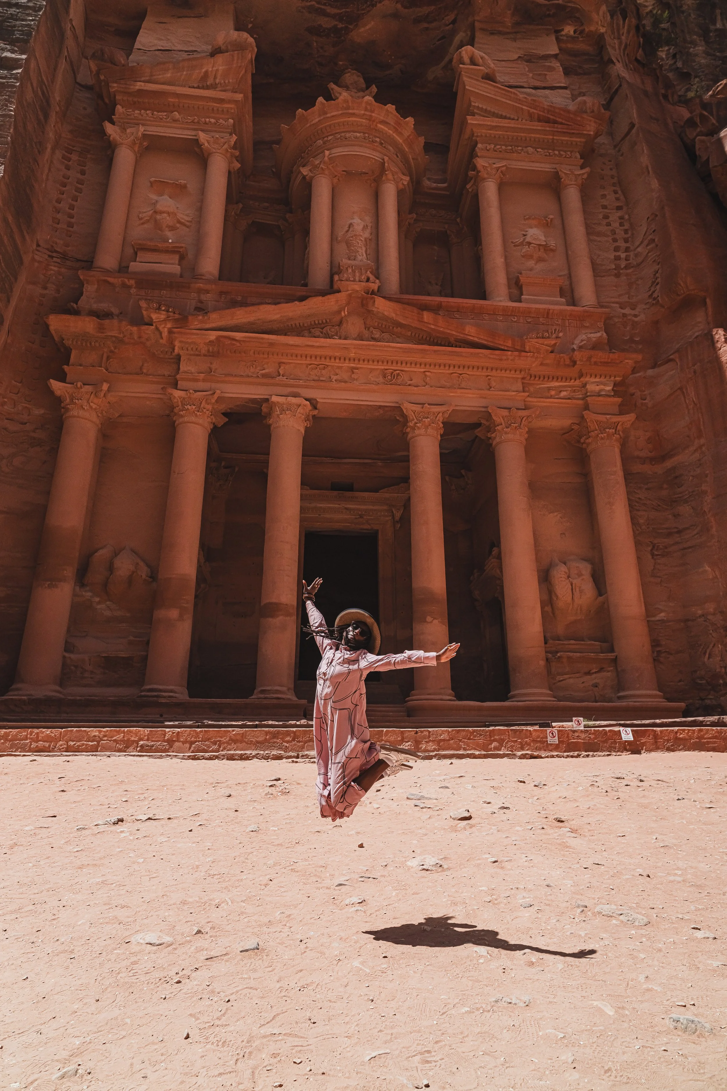 A woman in a long dress, sunglasses, and a wide-brimmed hat is jumping in front of the ancient rock-cut monument, Al-Khazneh, also known as The Treasury, in Petra, Jordan.