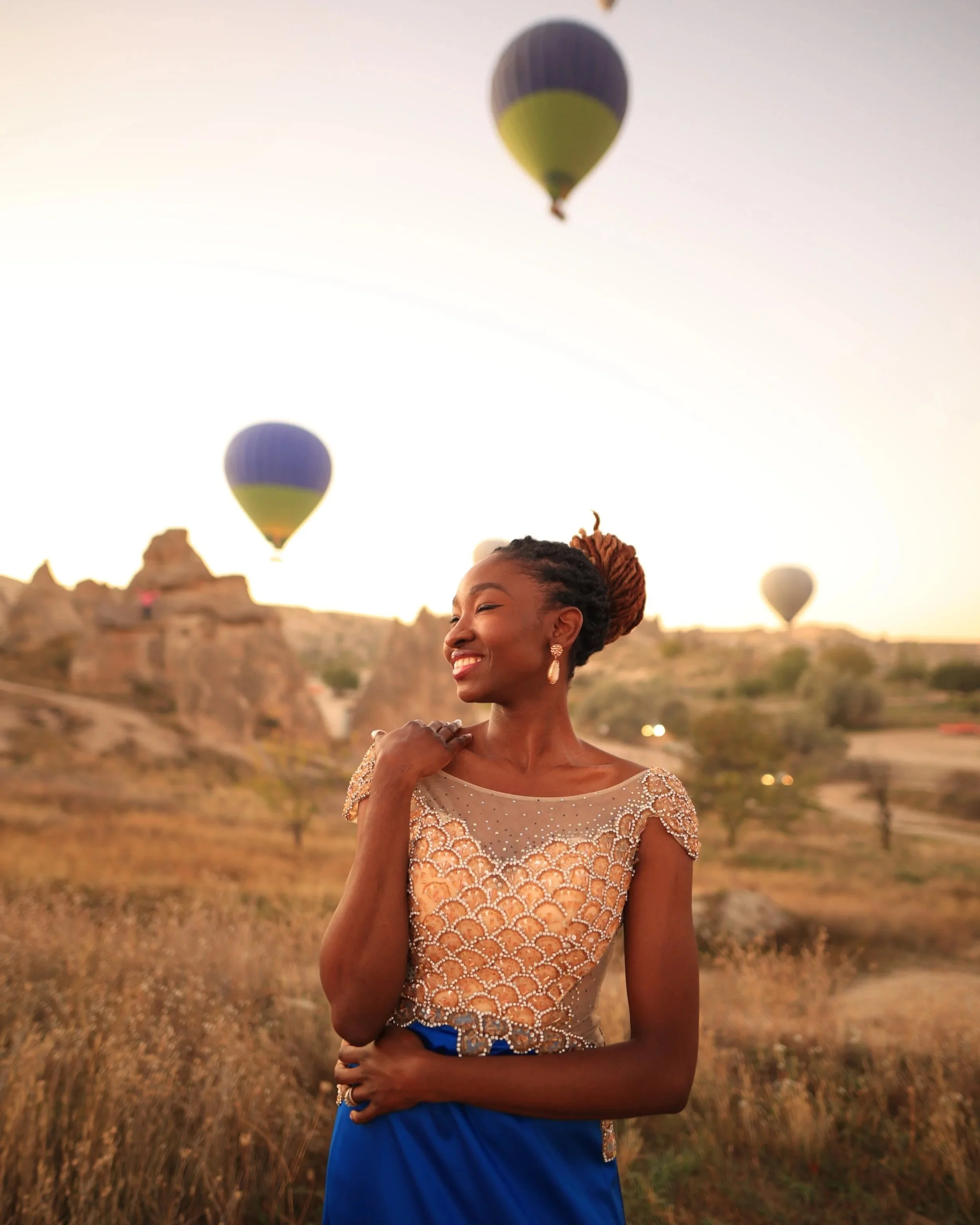 A woman with styled hair, earrings, and a beaded top smiles while standing outdoors at sunset with hot air balloons in the sky behind her.