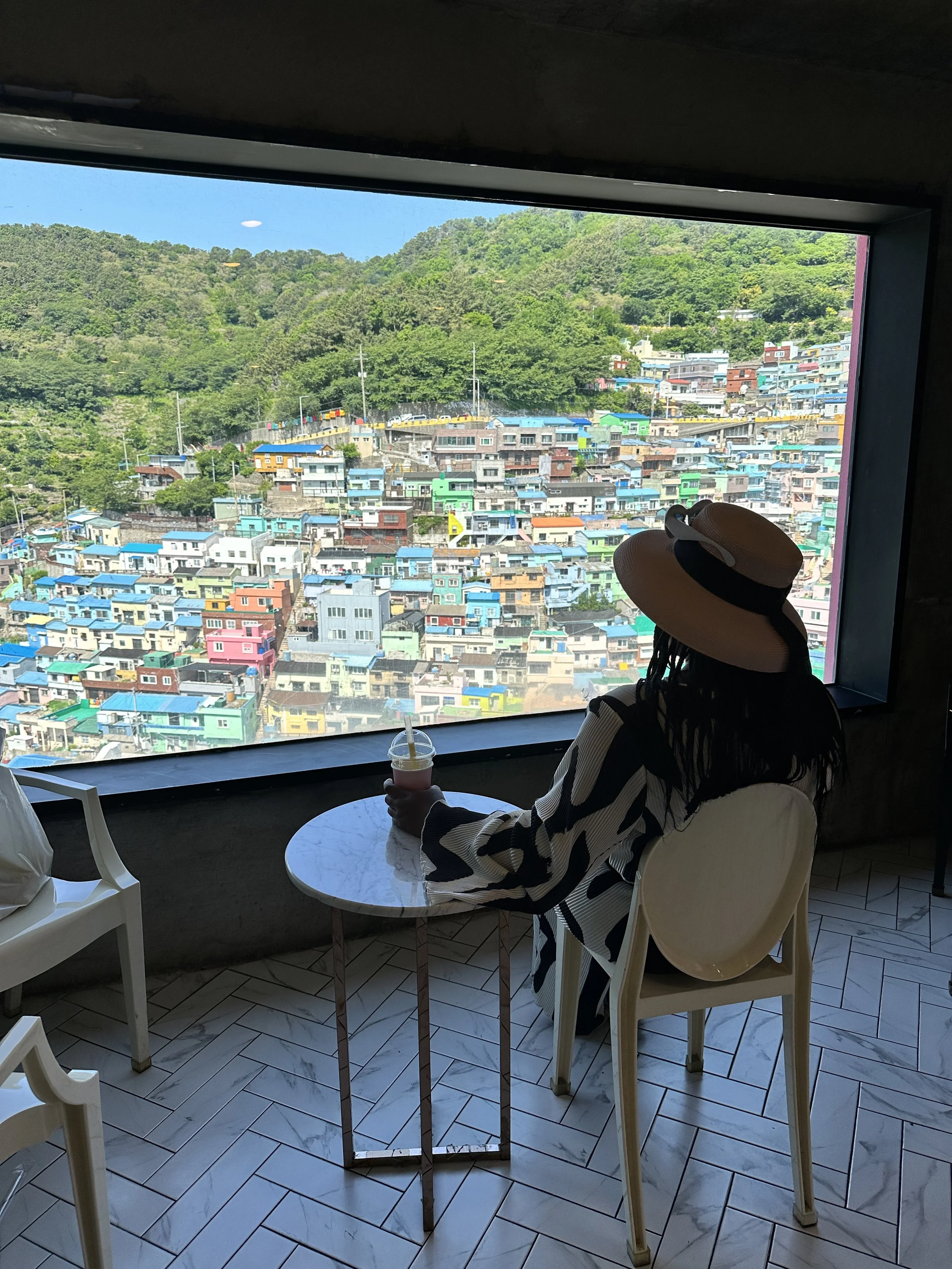 Person sitting at a small table inside a building, facing a large window with a view of colorful hillside houses and green trees, holding a drink.