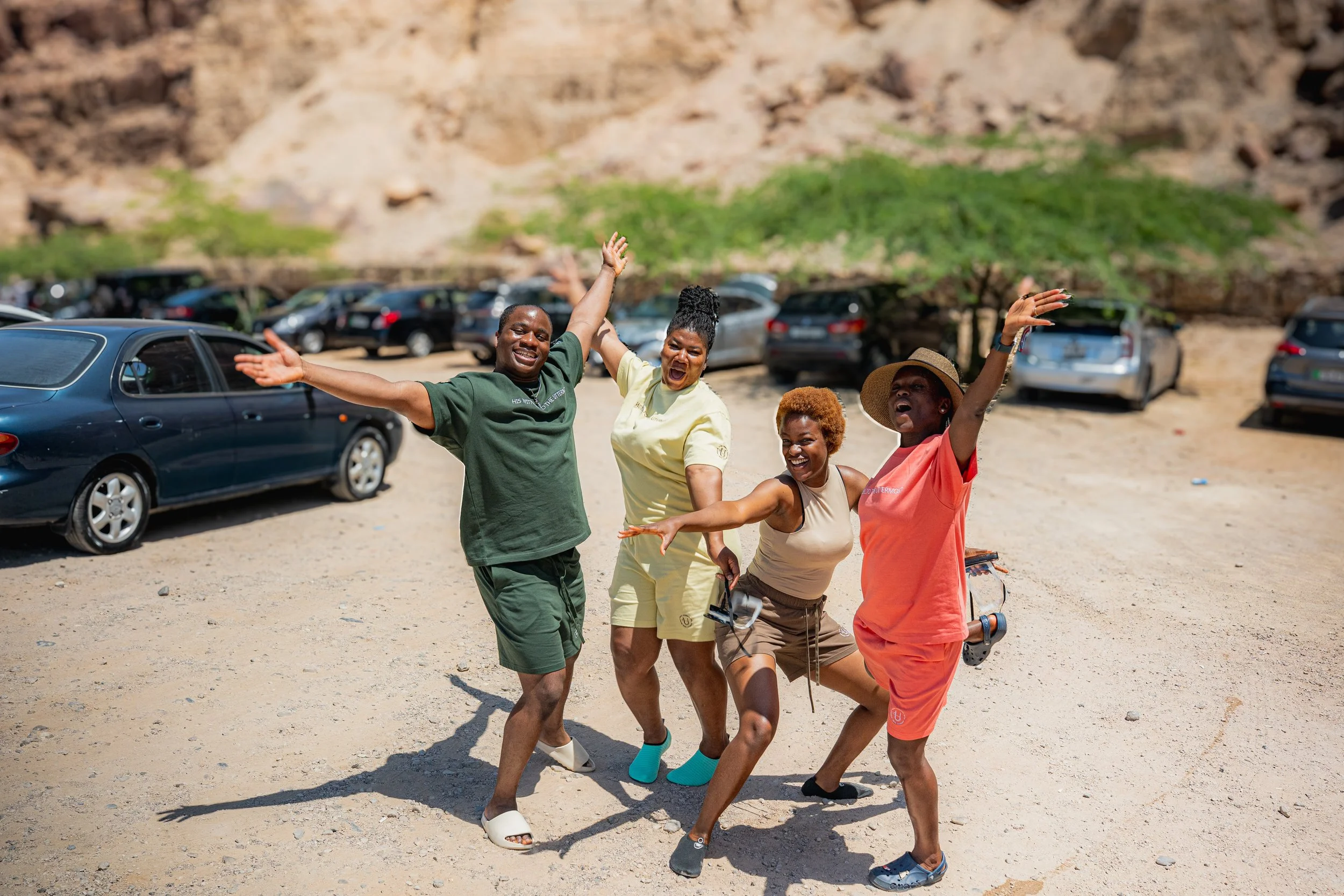 Four friends having fun and dancing in a parking lot near a rocky hillside. They are smiling and laughing.