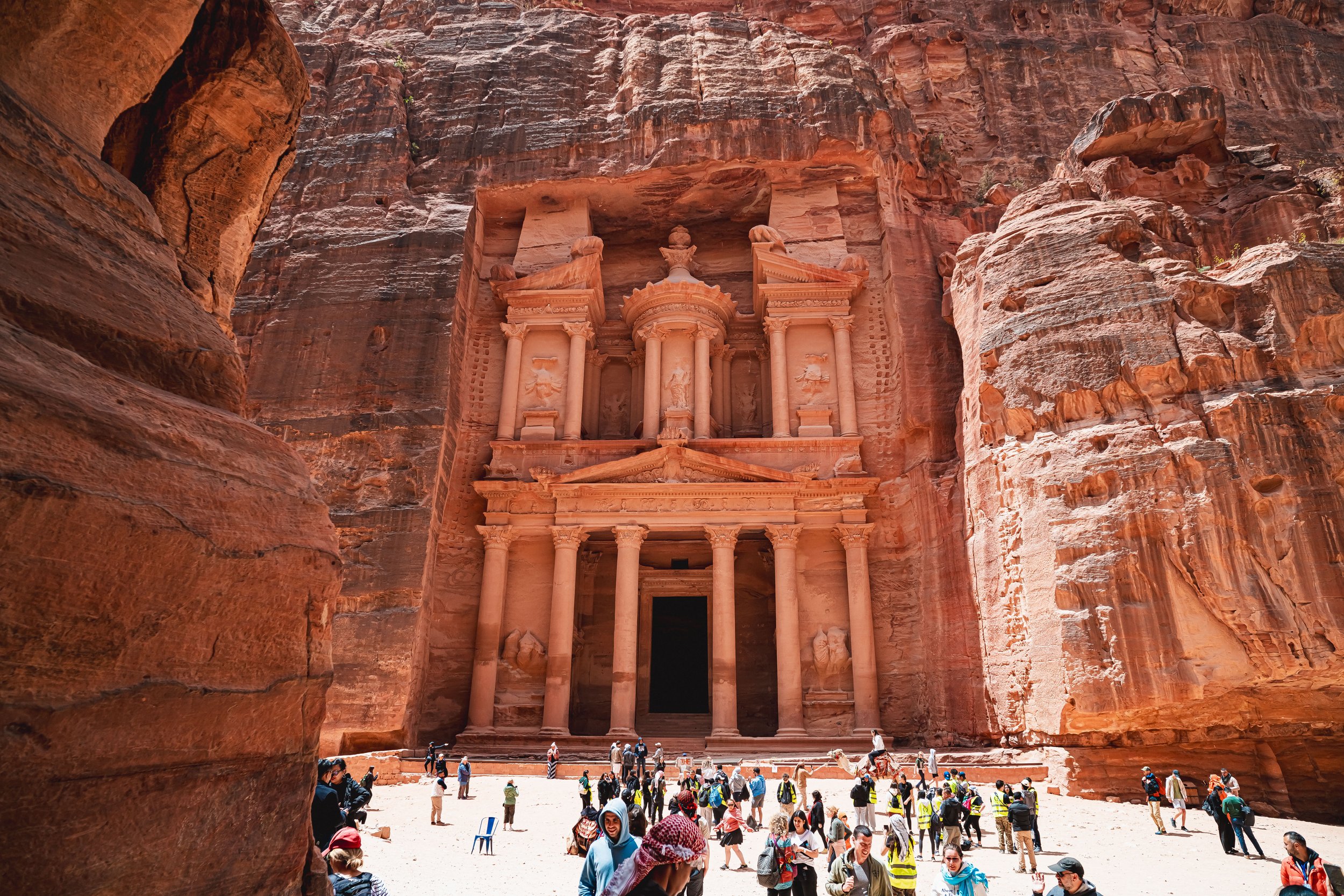 The image shows the ancient city of Petra, with the famous Treasury building carved into rose-red sandstone cliffs, filled with tourists exploring the site.