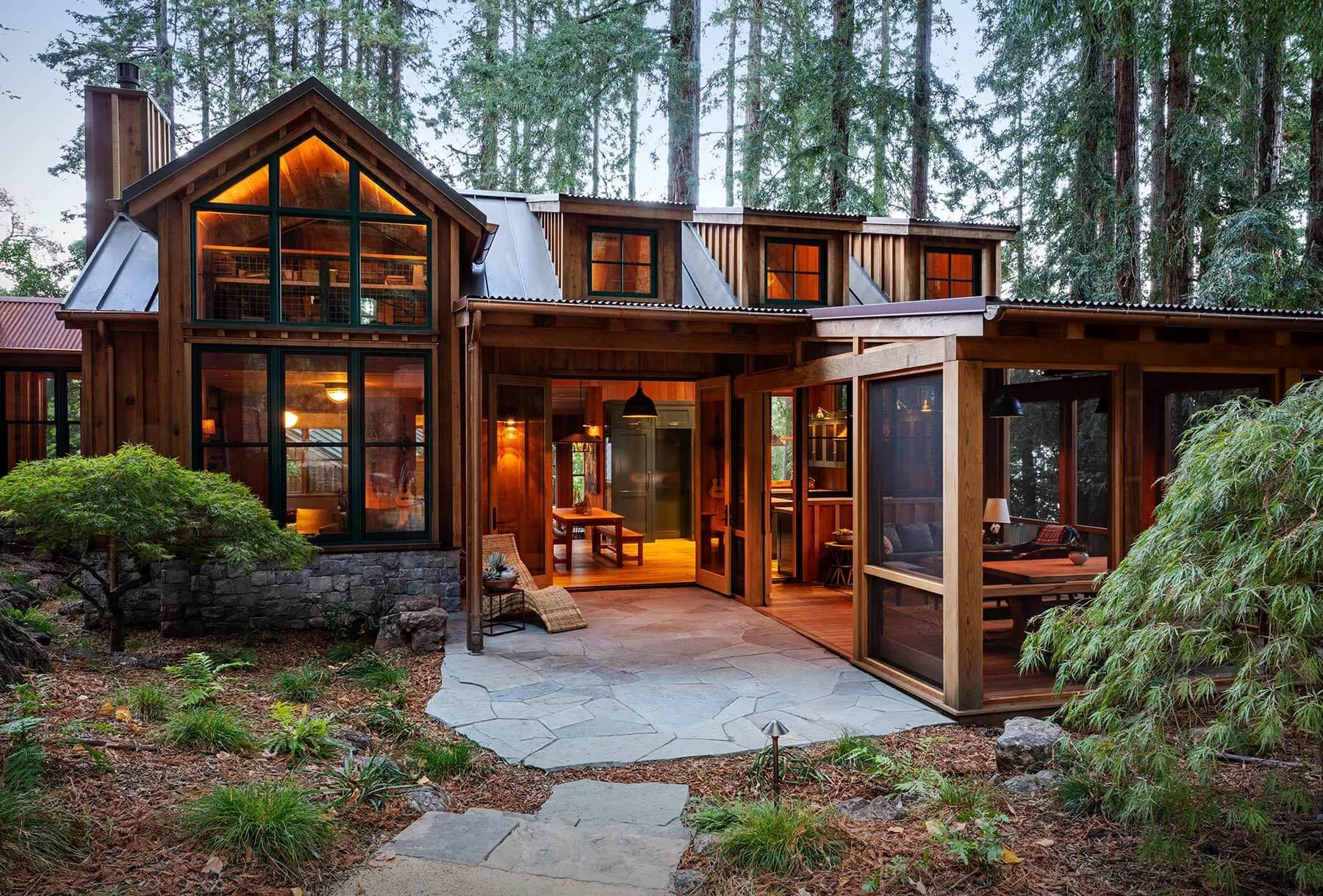 A multi-story wooden house with large glass windows, surrounded by tall trees in a forest. The house features a patio with outdoor seating, a stone pathway, and warm interior lighting.