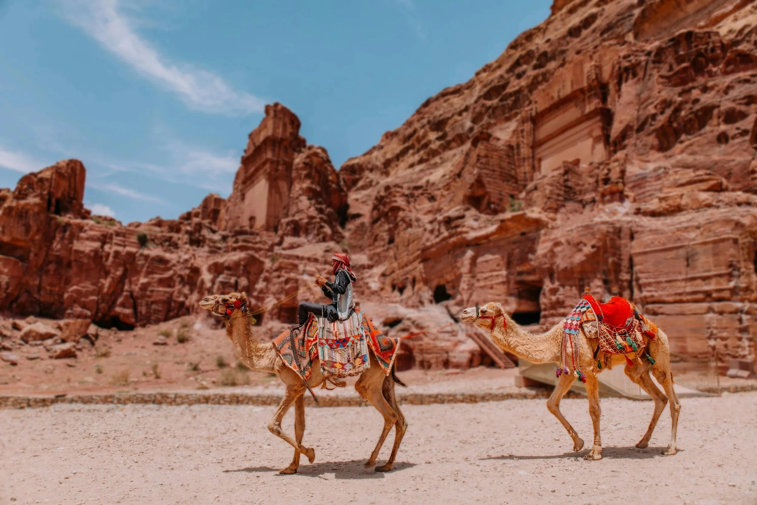 A person riding a camel in front of rocky desert cliffs under a blue sky, with another camel nearby.
