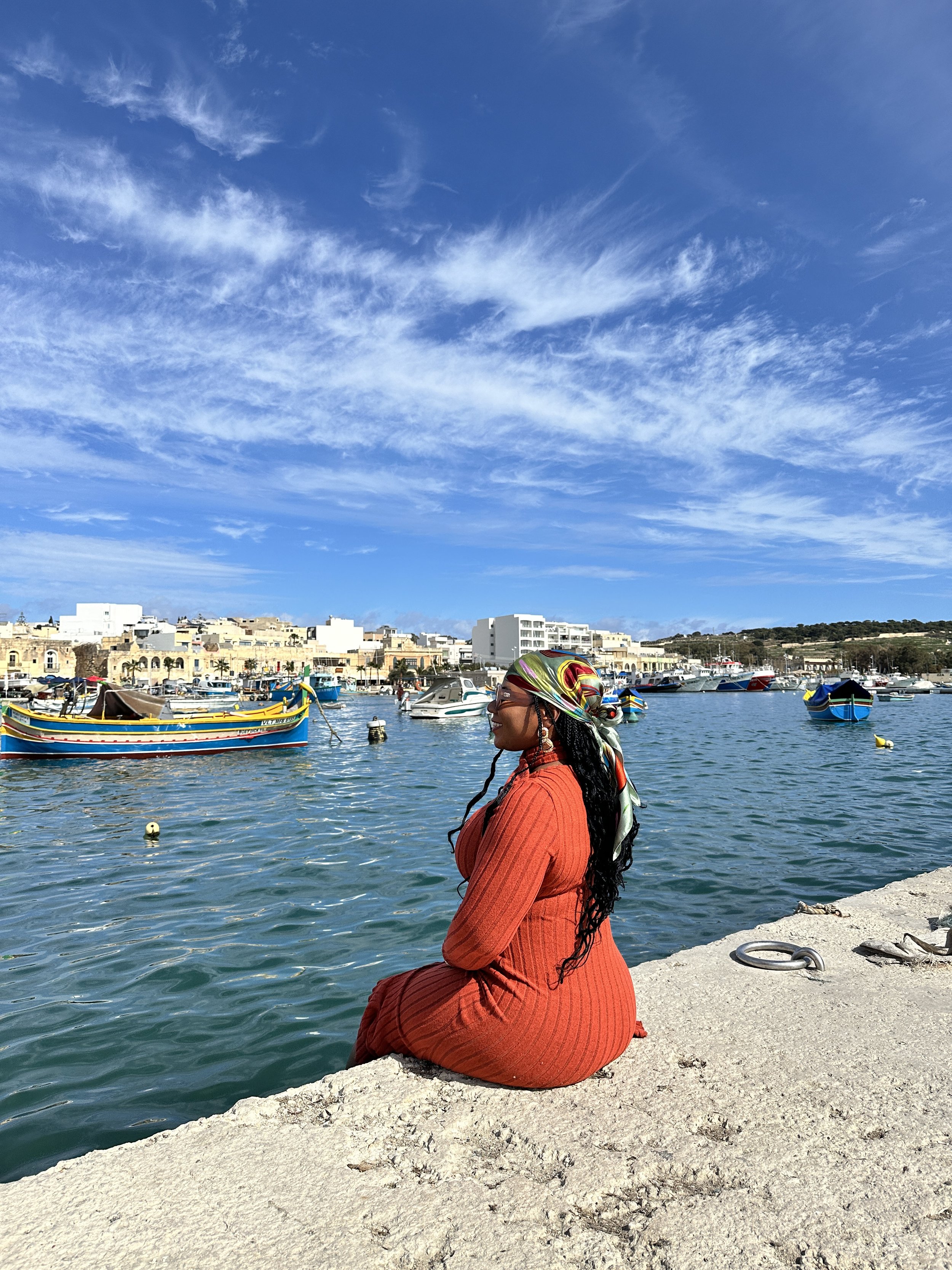 A woman sitting on a concrete dock beside a harbor with boats, wearing an orange dress and a multicolored headscarf under a blue sky with scattered clouds.