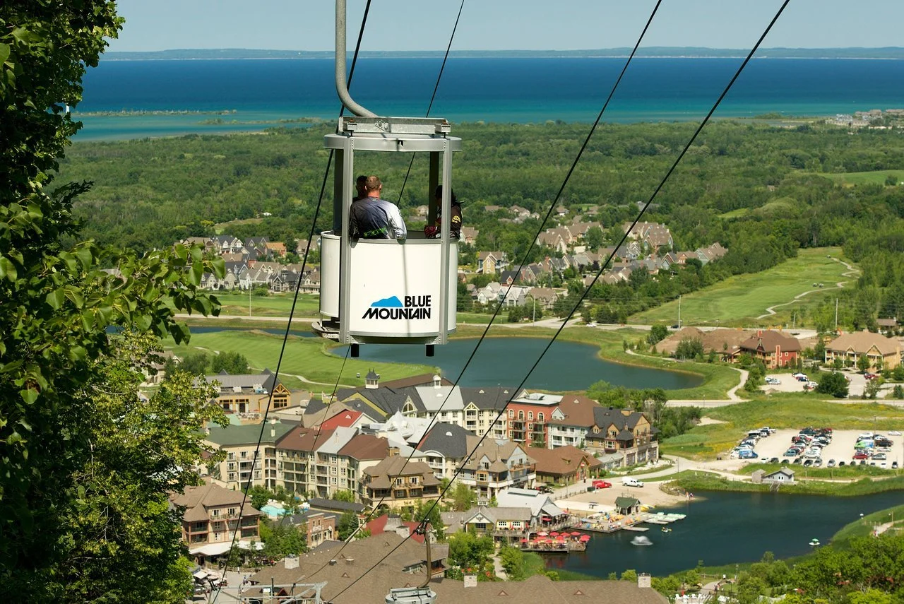 People riding a Blue Mountain chairlift above a town with lakes, green fields, and a view of a distant lake or ocean in the background.