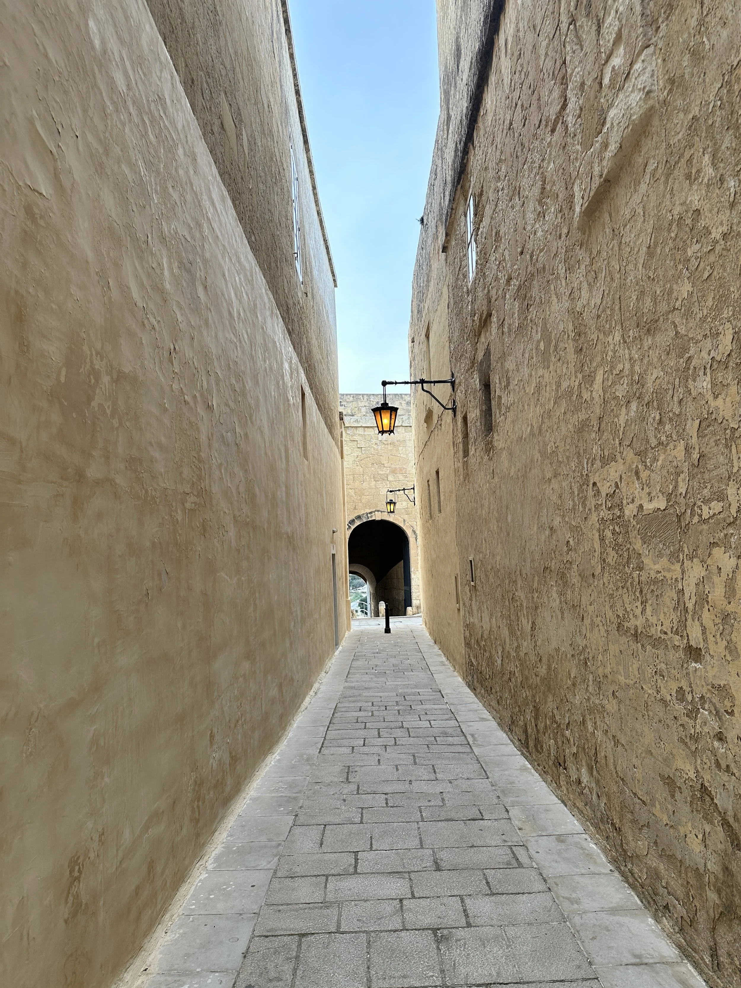 Narrow cobblestone alleyway with beige stucco walls and vintage lanterns, leading to a stone archway at the end under a blue sky.