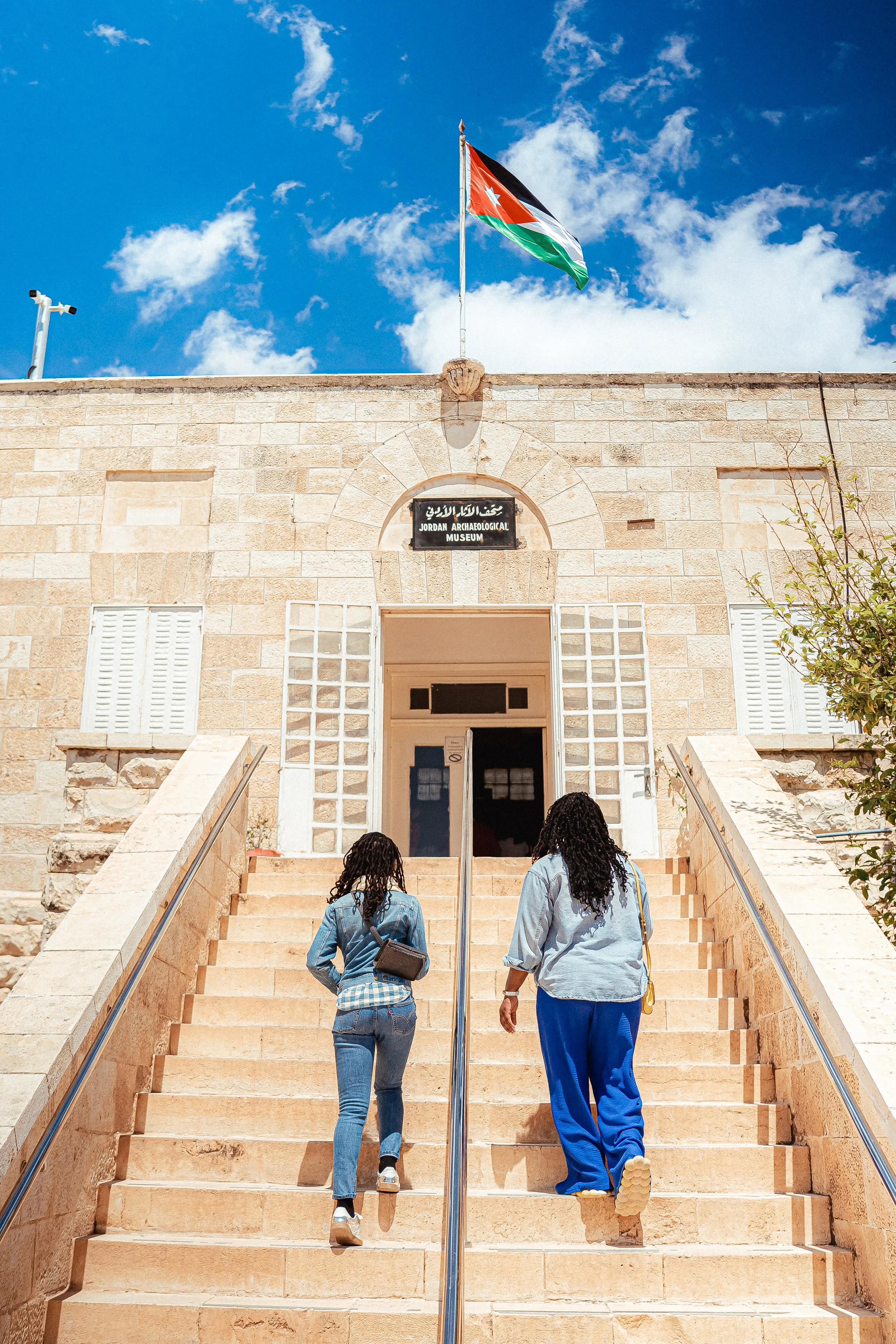 Two women climb stairs towards the Jordan Archaeological Museum, which has a sign and is topped with a Jordanian flag, under a partly cloudy sky.