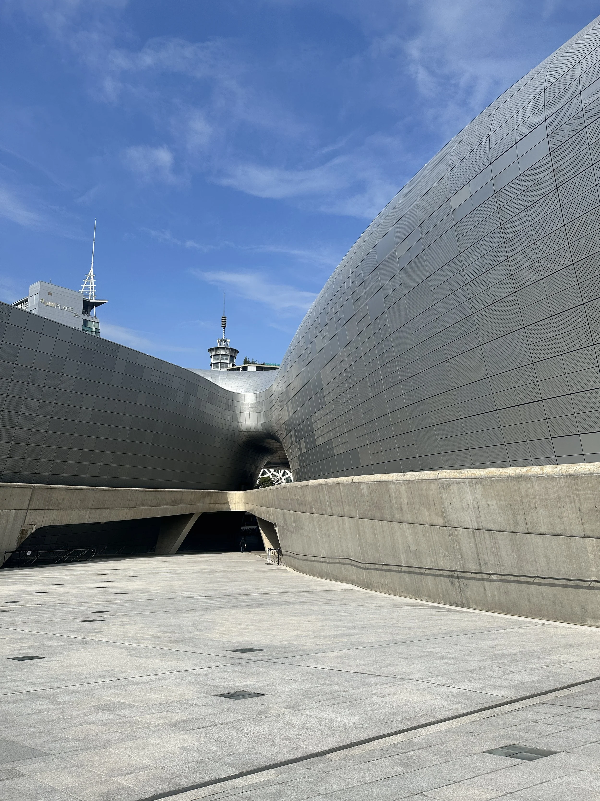 Modern architectural building with curved gray exterior walls and a wide open walkway beneath, set against a bright blue sky with scattered clouds.