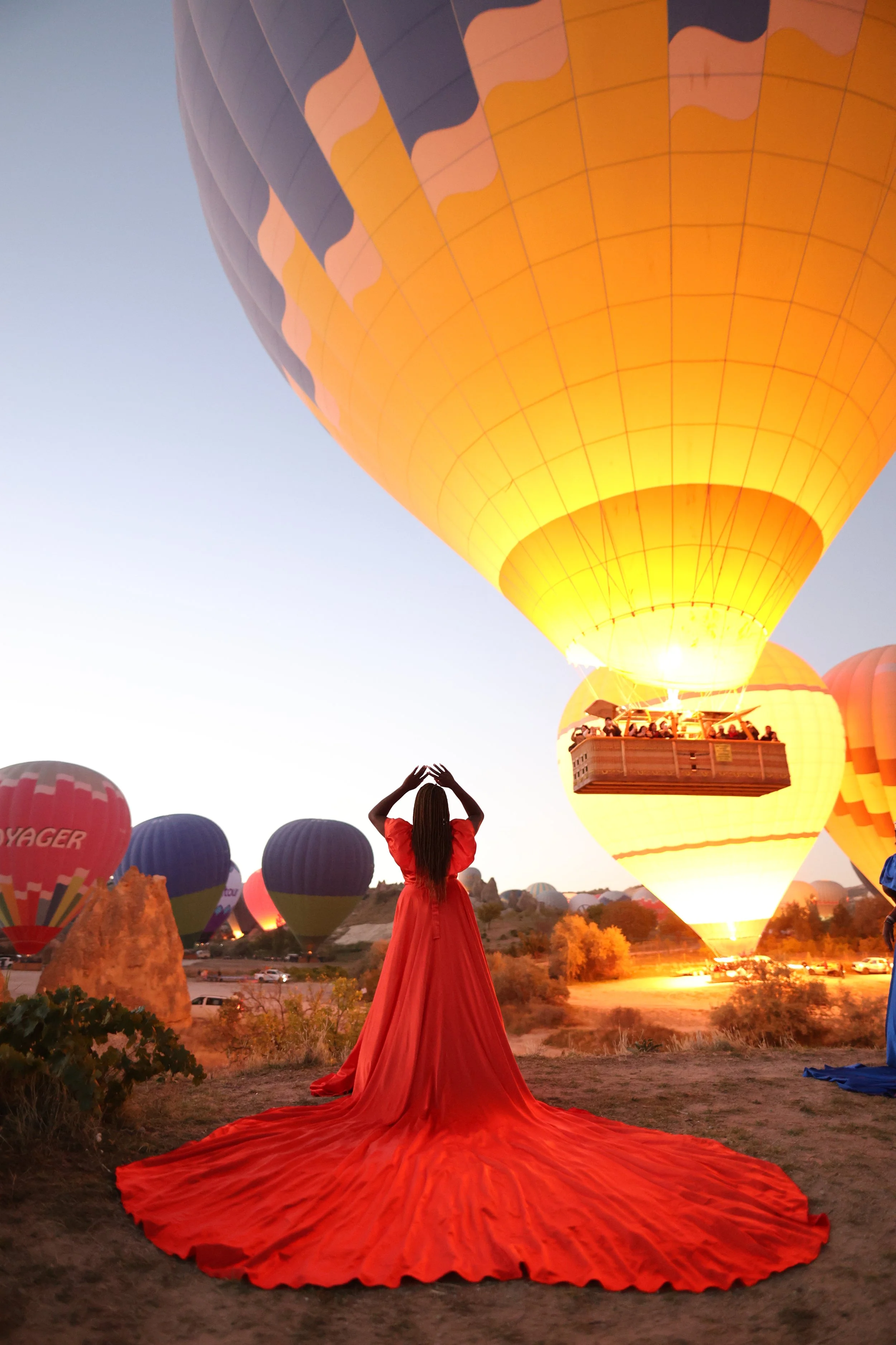 A woman in a long red dress stands on the ground with her back to the camera, watching hot air balloons glowing at sunrise or sunset, with several other balloons in the background.