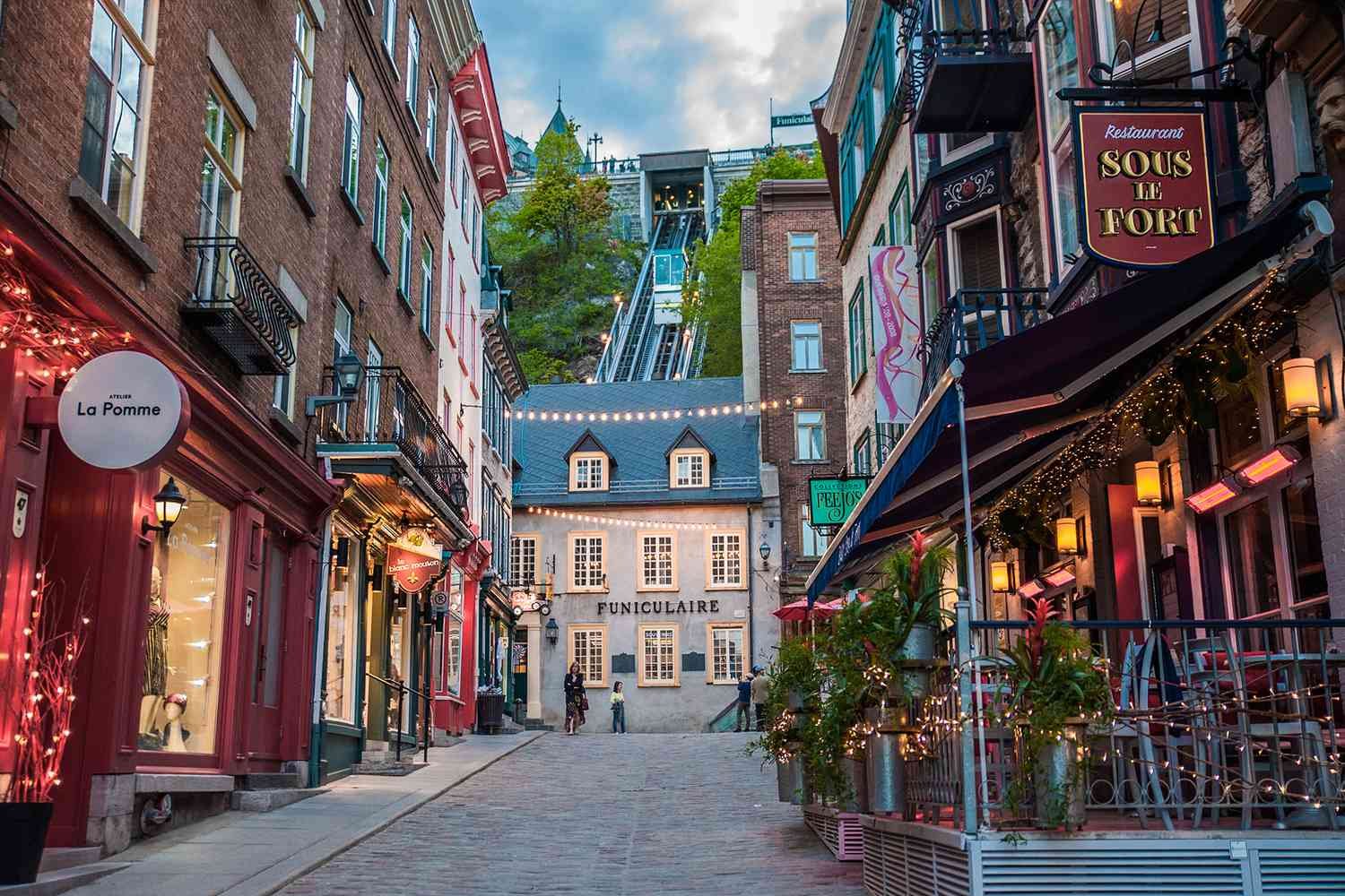 A narrow cobblestone street lined with colorful buildings, shops, and restaurants, leading up to Funiculaire in Montreal, with a funicular railway ascending a hill in the background.