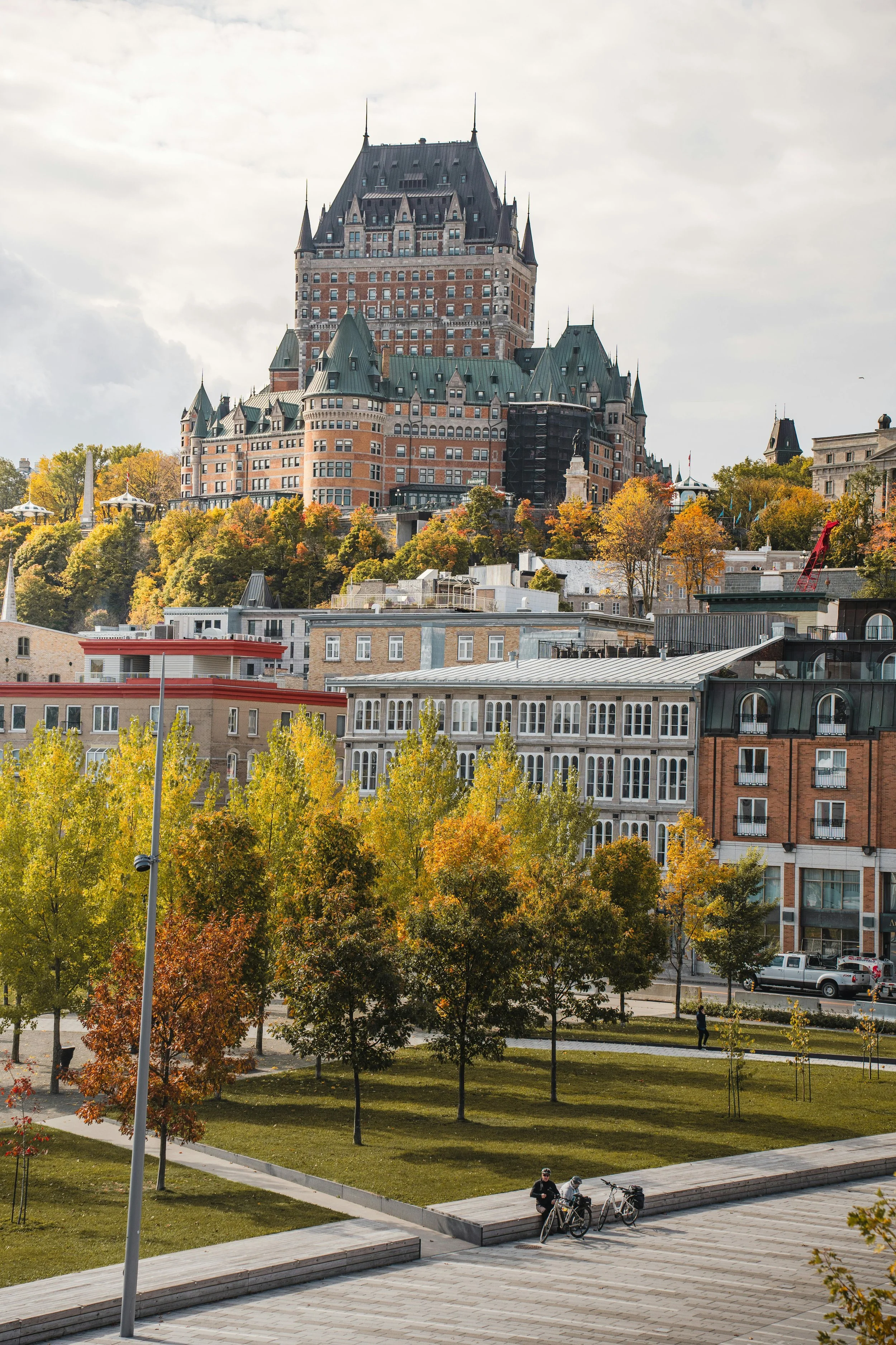 A cityscape featuring a historic hotel on a hill with colorful fall foliage, modern buildings, a park with trees, and people with bicycles in the foreground.