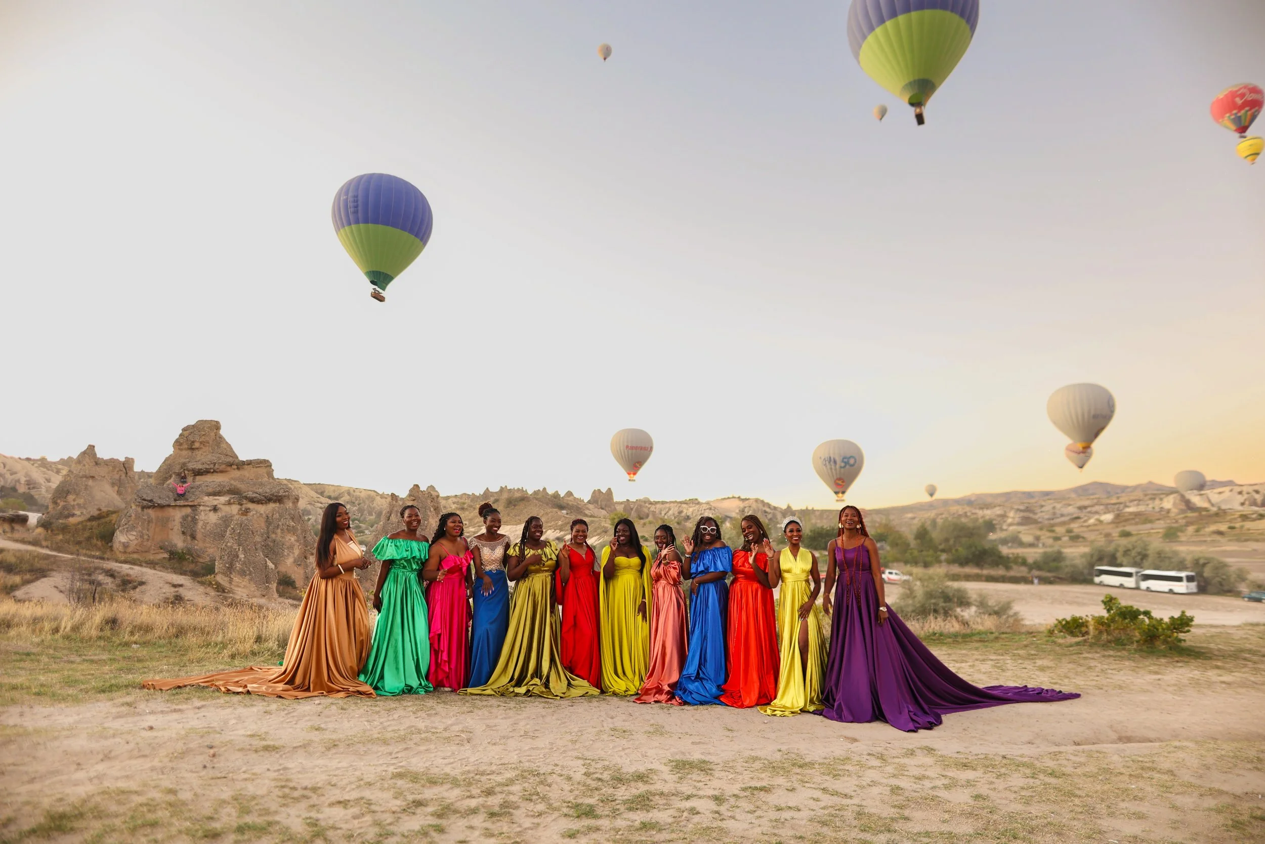 Group of women in colorful dresses standing outdoors with hot air balloons in the sky.