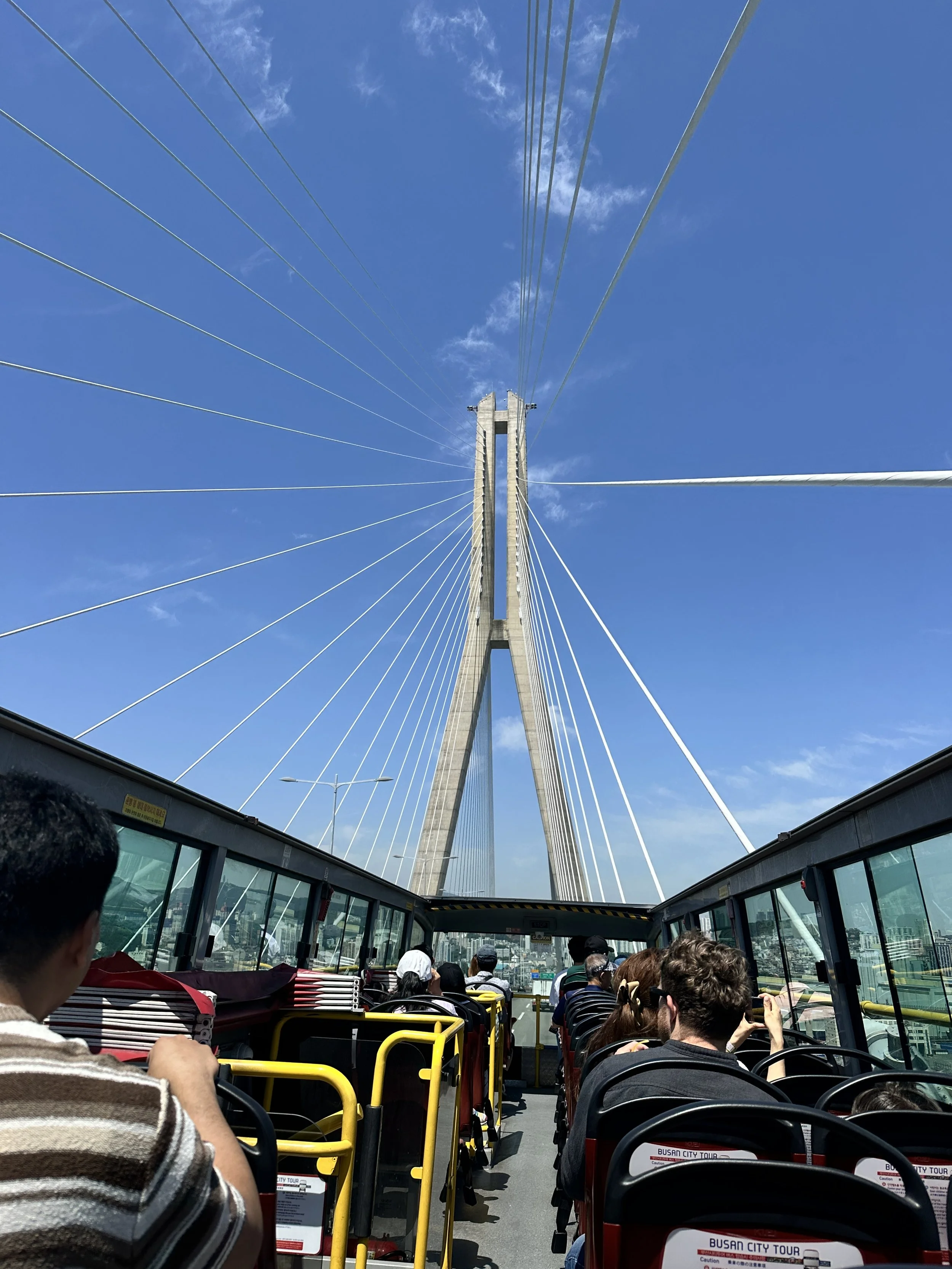Tour bus with passengers riding towards a large cable-stayed bridge on a sunny day.