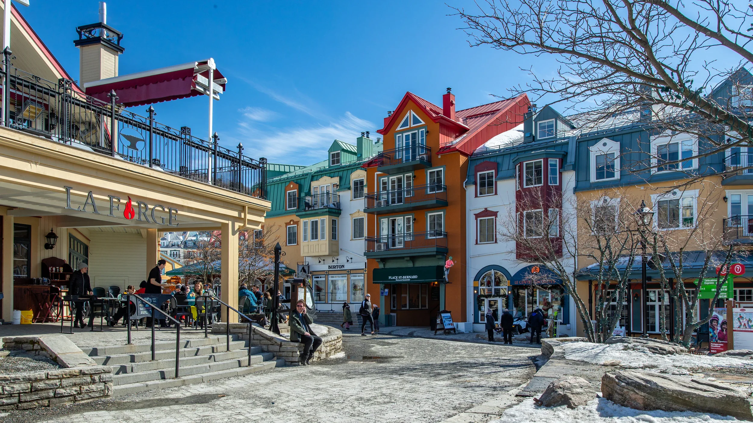 Colorful buildings with cafes and shops on a street, some people sitting and walking outside, leafless trees, and clear blue sky.