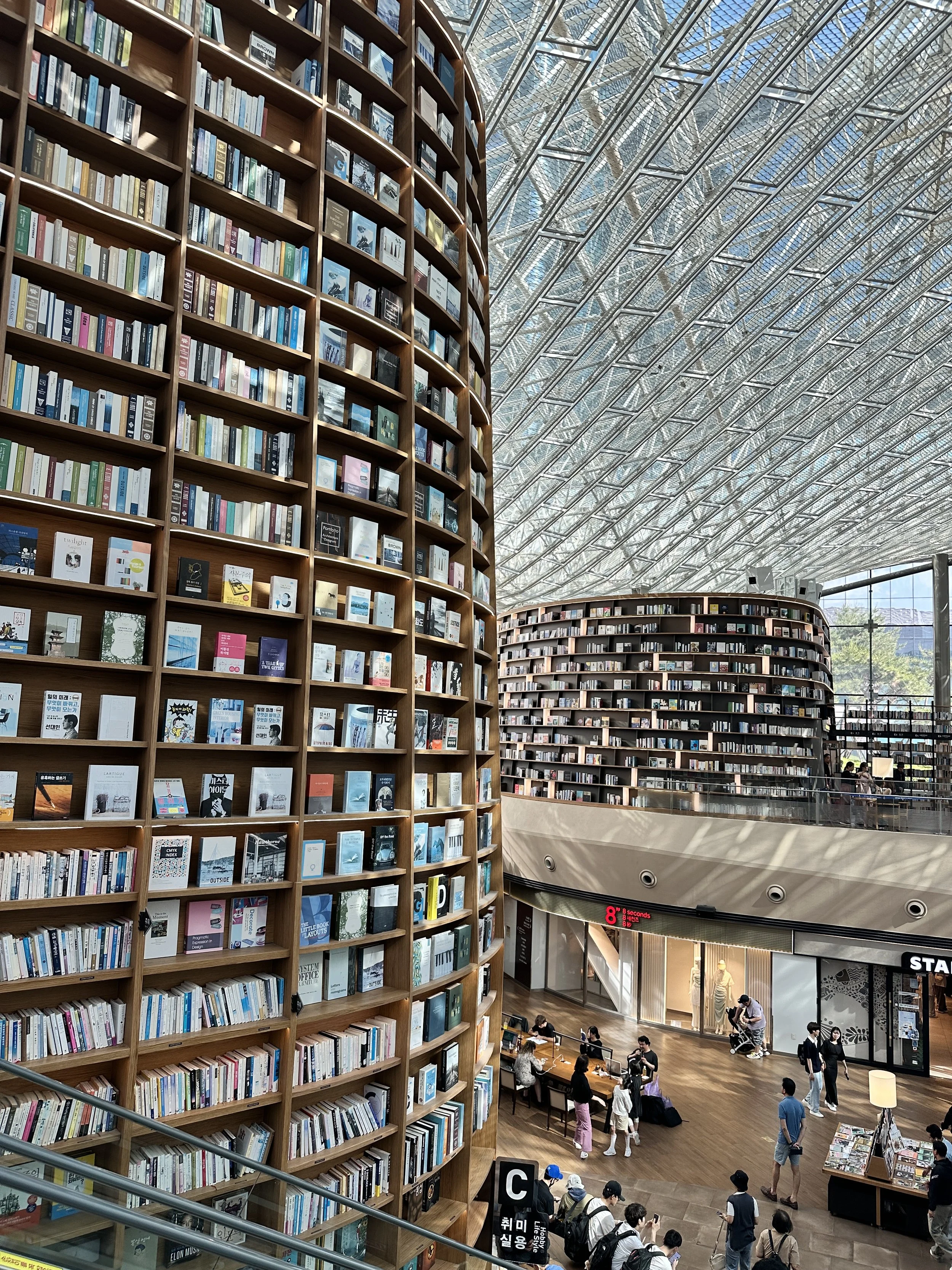 Inside a modern library with tall, curved wooden bookshelves filled with books. The ceiling is made of glass panels allowing natural light. People are walking and sitting at tables.