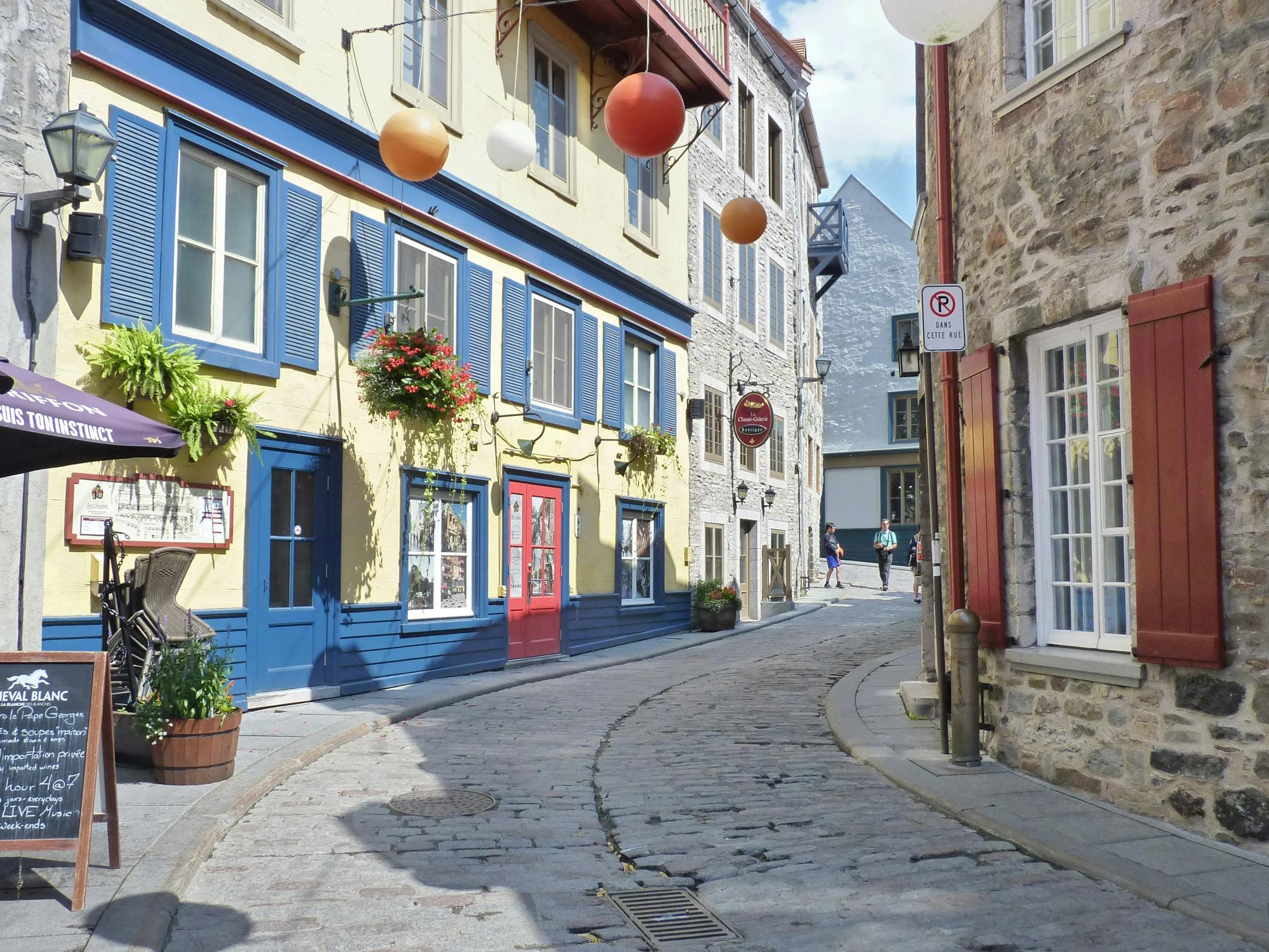 A narrow cobblestone street in a Québec City with colorful buildings, hanging flower baskets, and pedestrians walking in the distance under a partly cloudy sky.