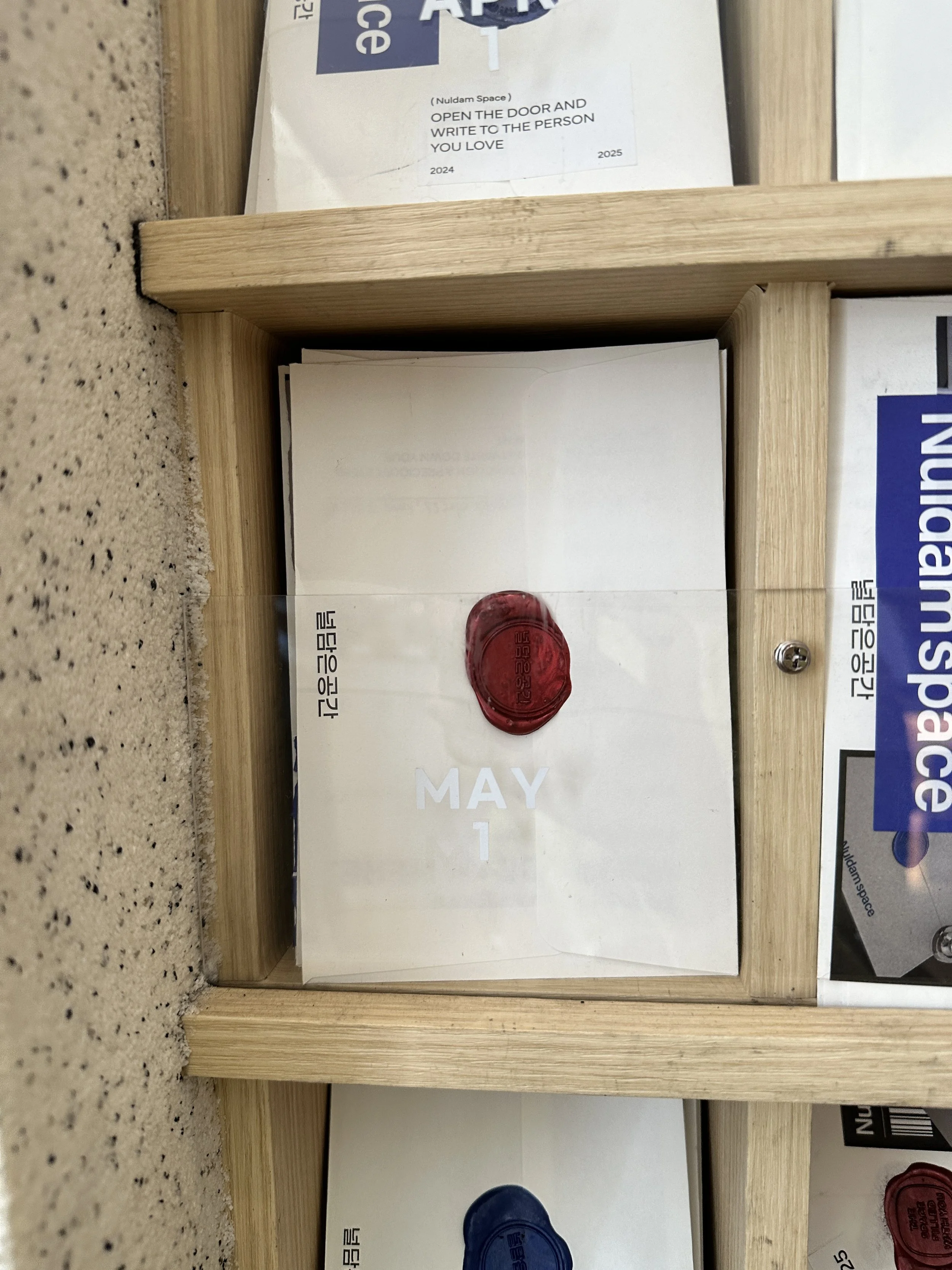 A letter with a red wax seal and the date 'May 1' on it, displayed in a wooden compartment with other similar letters and notices.