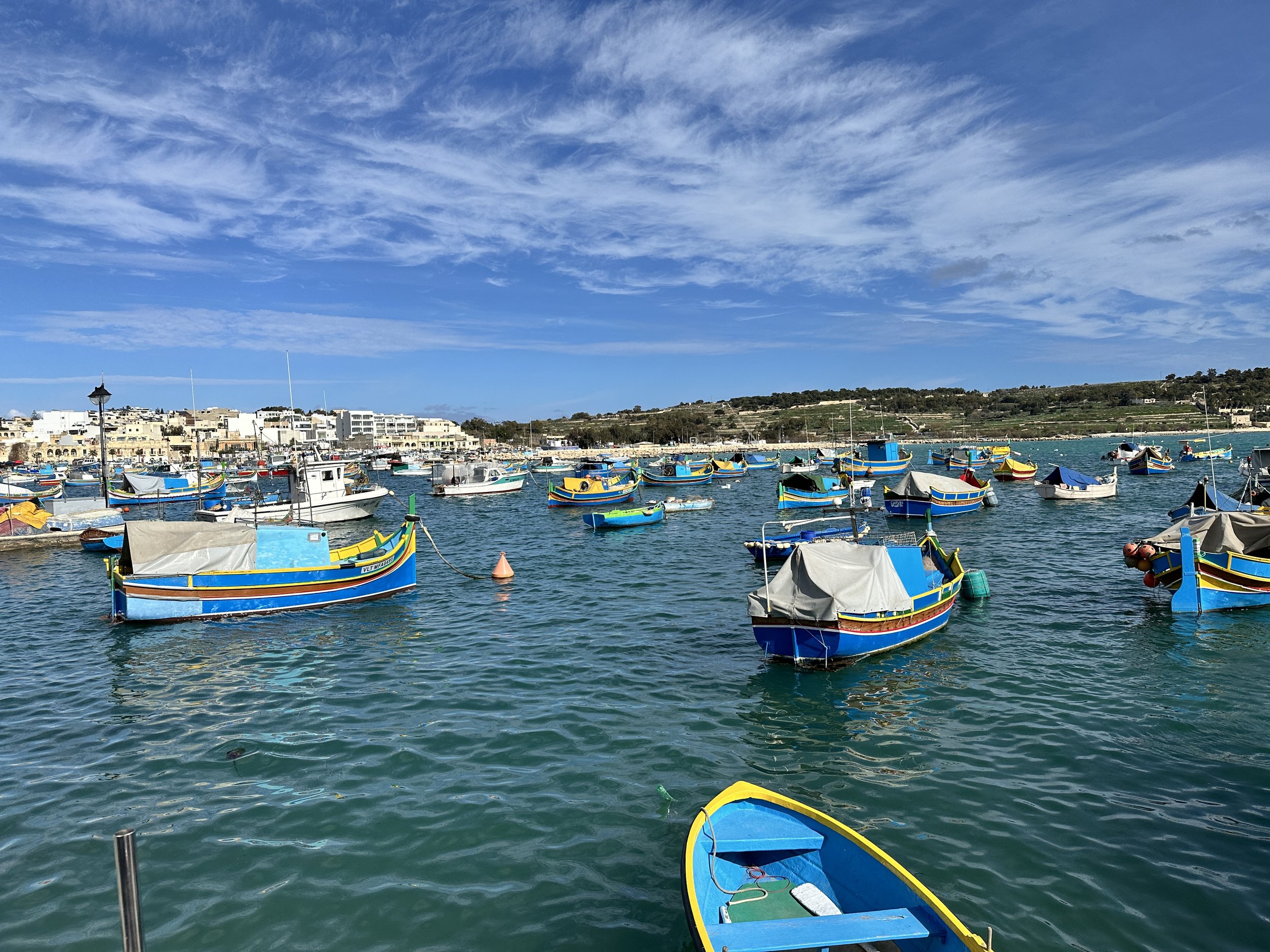 A harbor filled with colorful boats on calm waters, with a town and hills in the background under a partly cloudy blue sky.