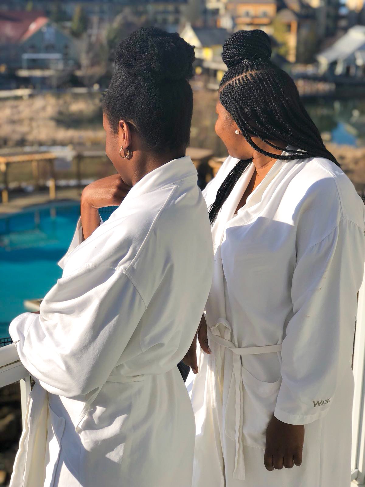 Two women dressed in white robes are standing outdoors, looking away from the camera, with a body of water and residential houses in the background.