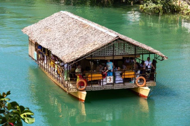 A floating house or restaurant with a thatched roof on a body of water, with people dining on the deck.
