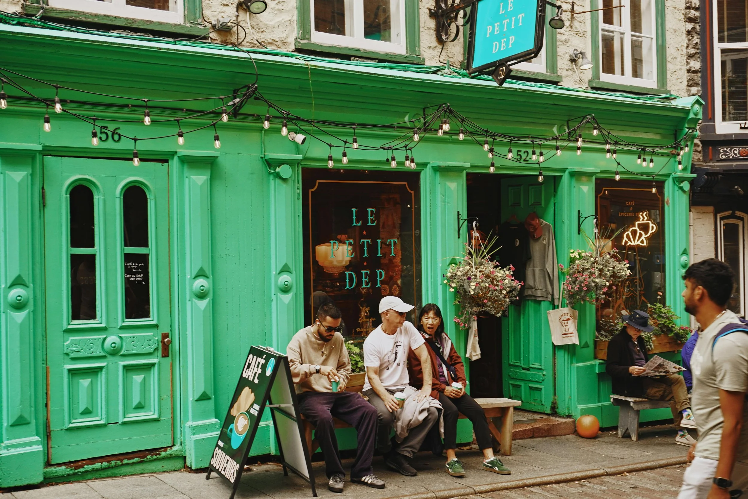 People sitting outside a vibrant green cafe storefront named 'Le Petit Dep' on a street in an urban area