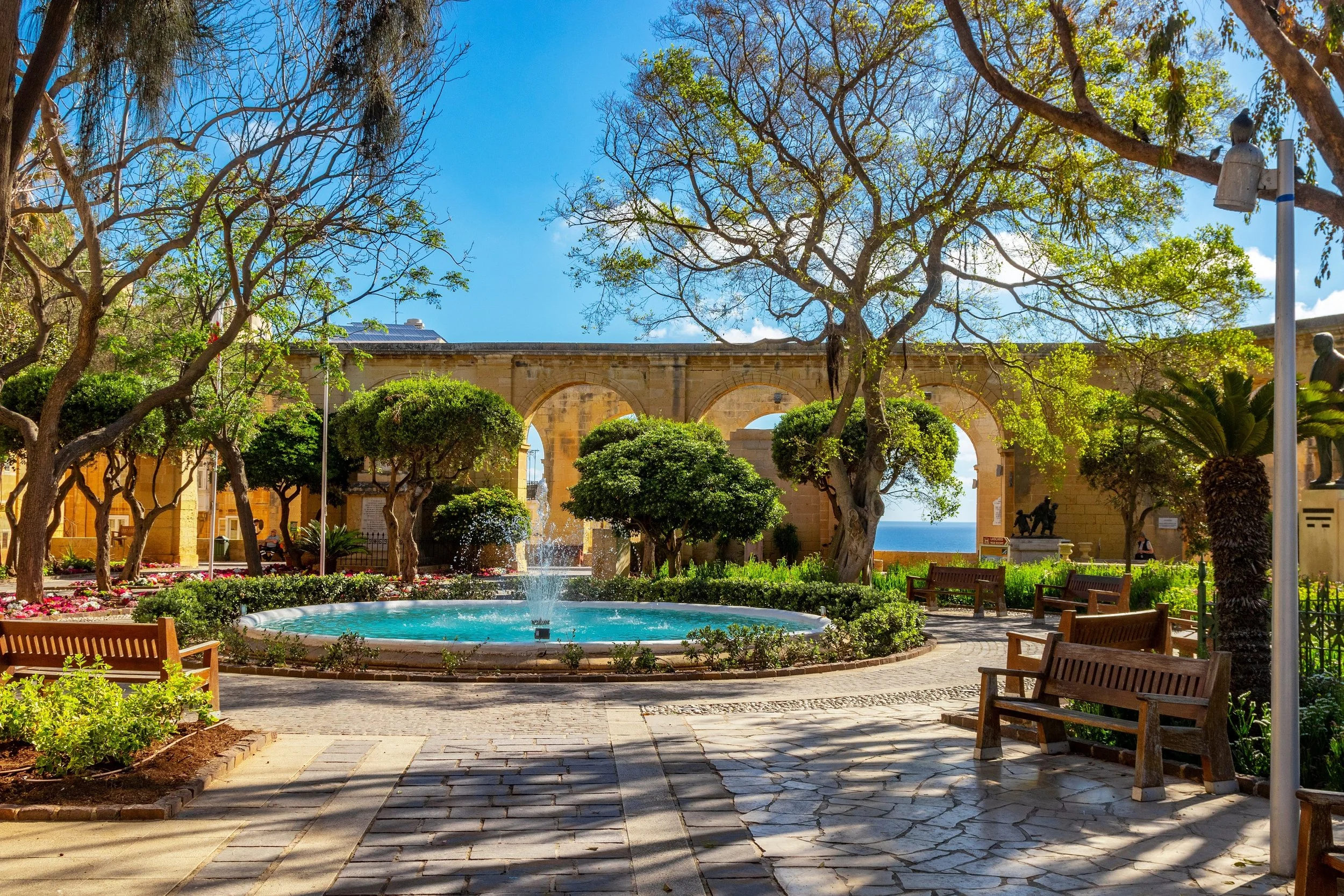 A park with a circular fountain in the center, surrounded by trees and benches. An ancient stone aqueduct with arches is visible in the background, along with a clear blue sky.