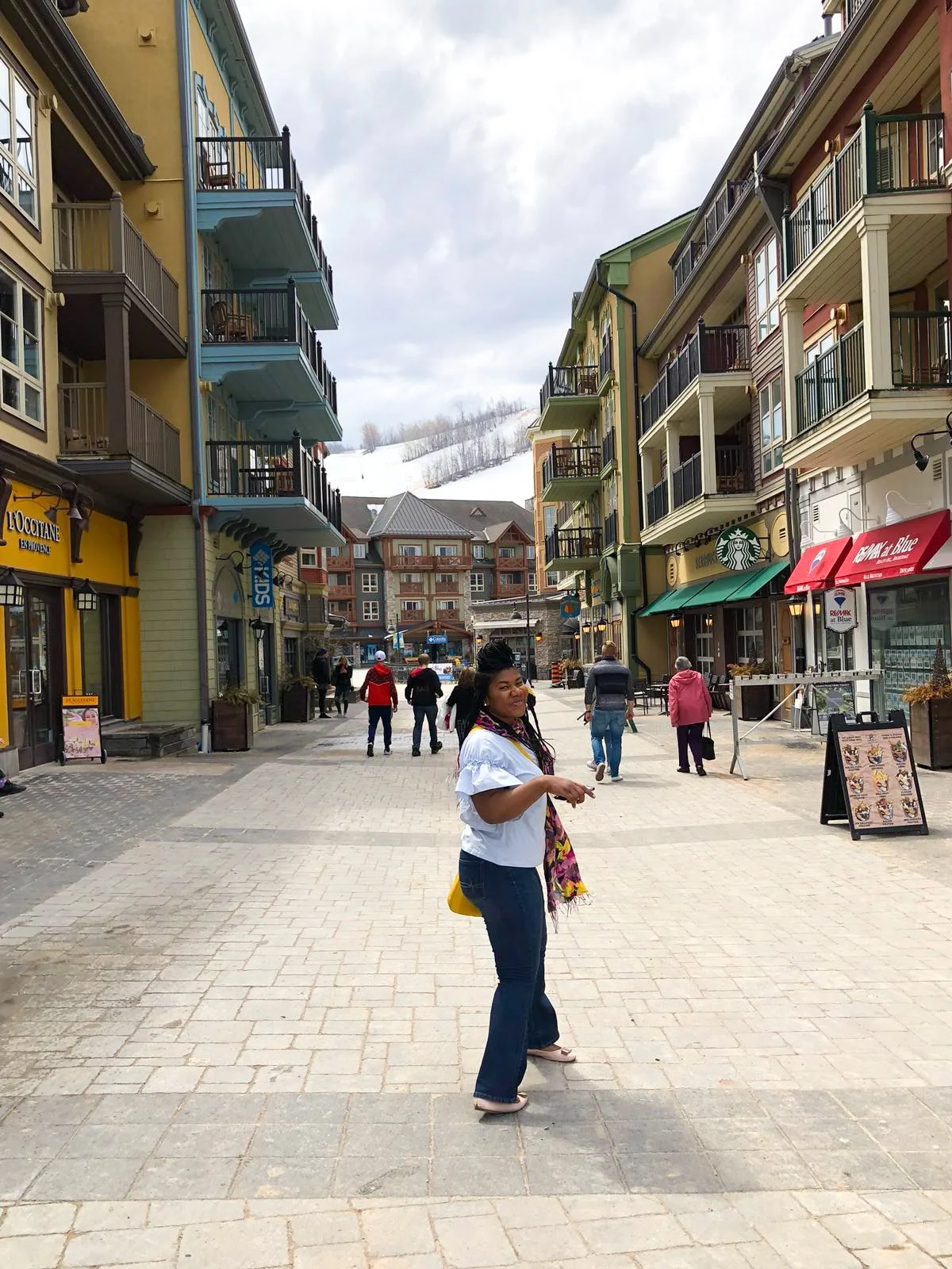 A woman standing in the middle of a pedestrian street in a mountain town with buildings on either side. Snow-capped mountains are visible in the background.