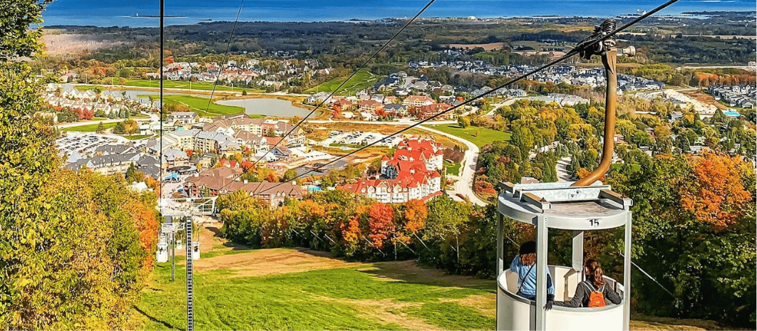 View from a cable car over a city surrounded by trees with fall foliage, a golf course, and distant hills.